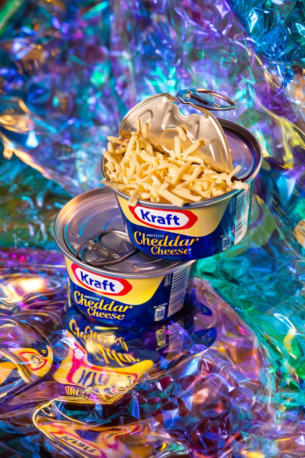 Two cans of Kraft processed cheddar cheese with one open can filled with shredded cheese, set against a colorful iridescent background.