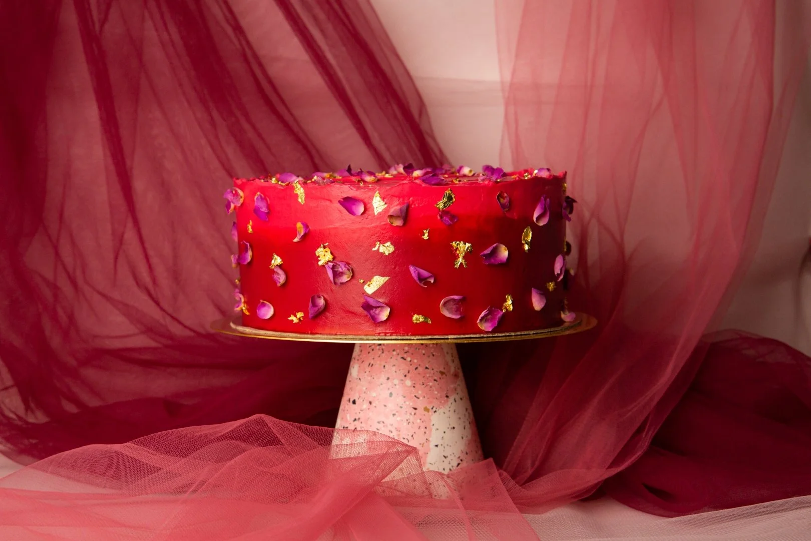 A red, round cake decorated with pink and purple flower petals and gold leaf, sitting on a speckled pink and white cake stand, with pink tulle fabric in the background.