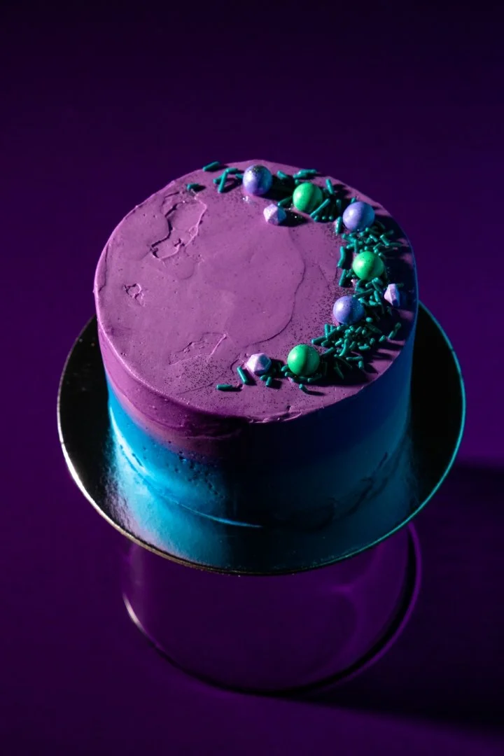 A round purple and blue cake with purple icing, decorated with purple, green, and silver spherical and sprinkles on top, on a reflective cake stand.