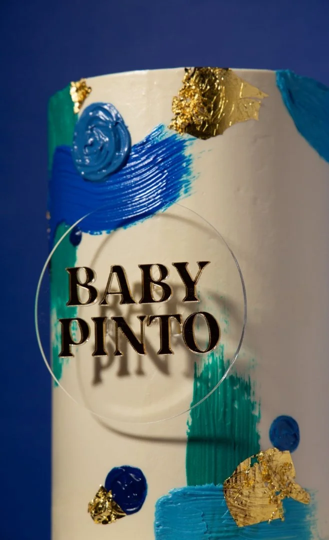 Close-up of a decorated baby shower cake with gold, blue, and green accents, and the words 'Baby Pinto' on the front.