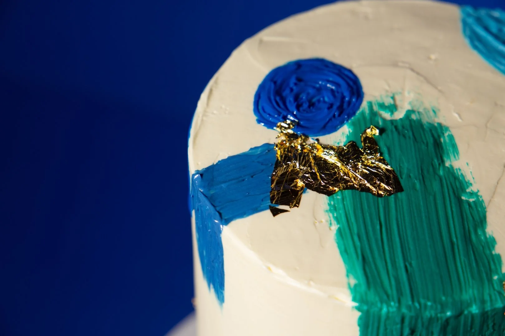 A cake decorated with abstract blue, green, and gold frosting on top, with a gold foil accent.