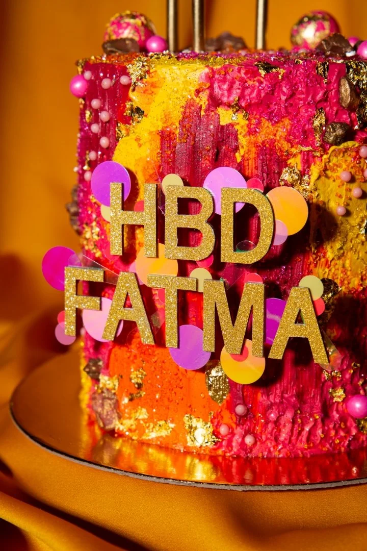 Colorful birthday cake with pink, yellow, and gold decoration, topped with chocolate and decorative spheres, with a gold "HBD FATMA" sign on it.