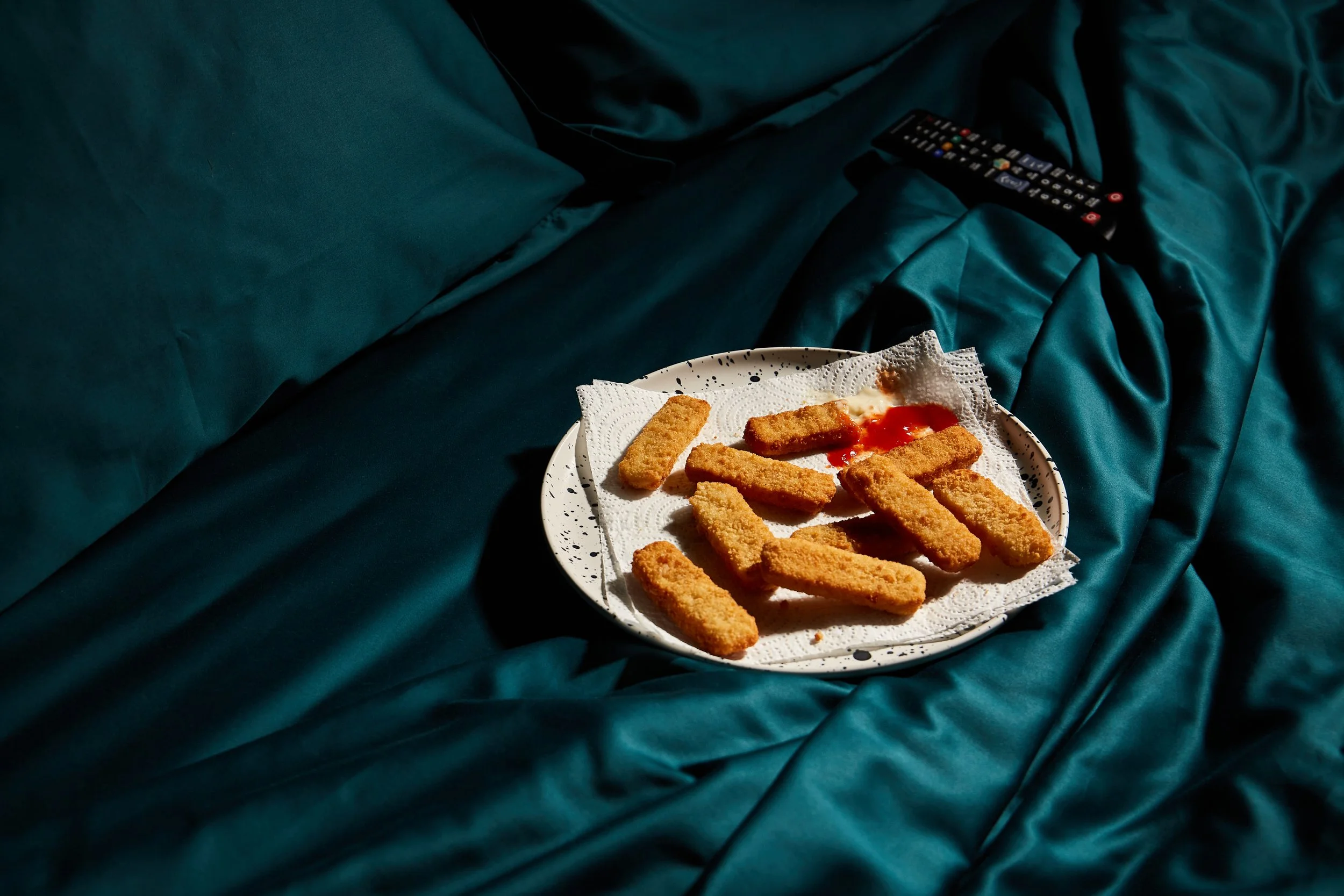 Plate of breaded chicken nuggets with ketchup on a white plate on teal bed with a remote control nearby.