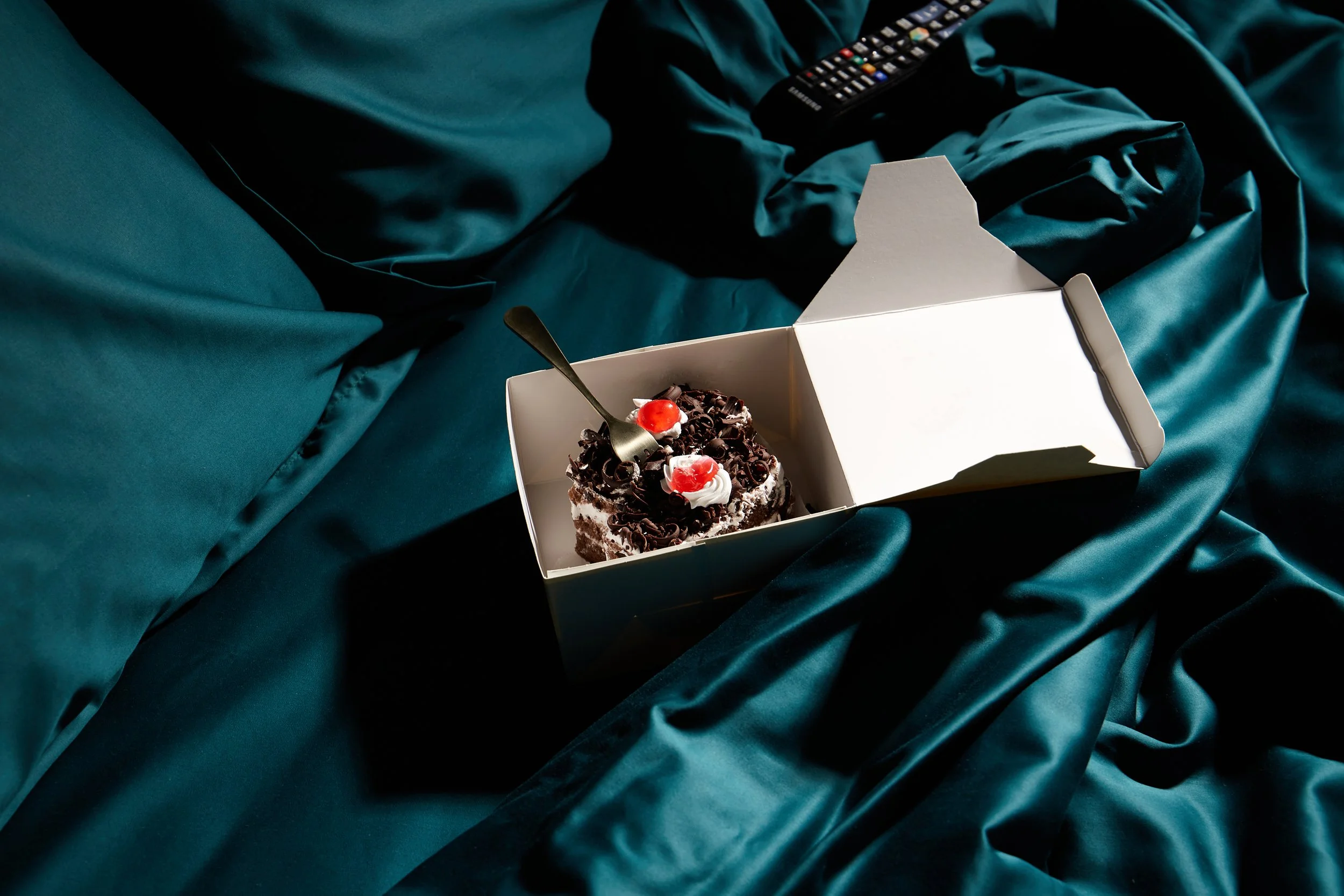 A partially opened white bakery box with a chocolate cake topped with whipped cream, cherries, and chocolate shavings inside on a dark teal satin bedsheet, with a spoon inserted into the cake and a TV remote in the background.