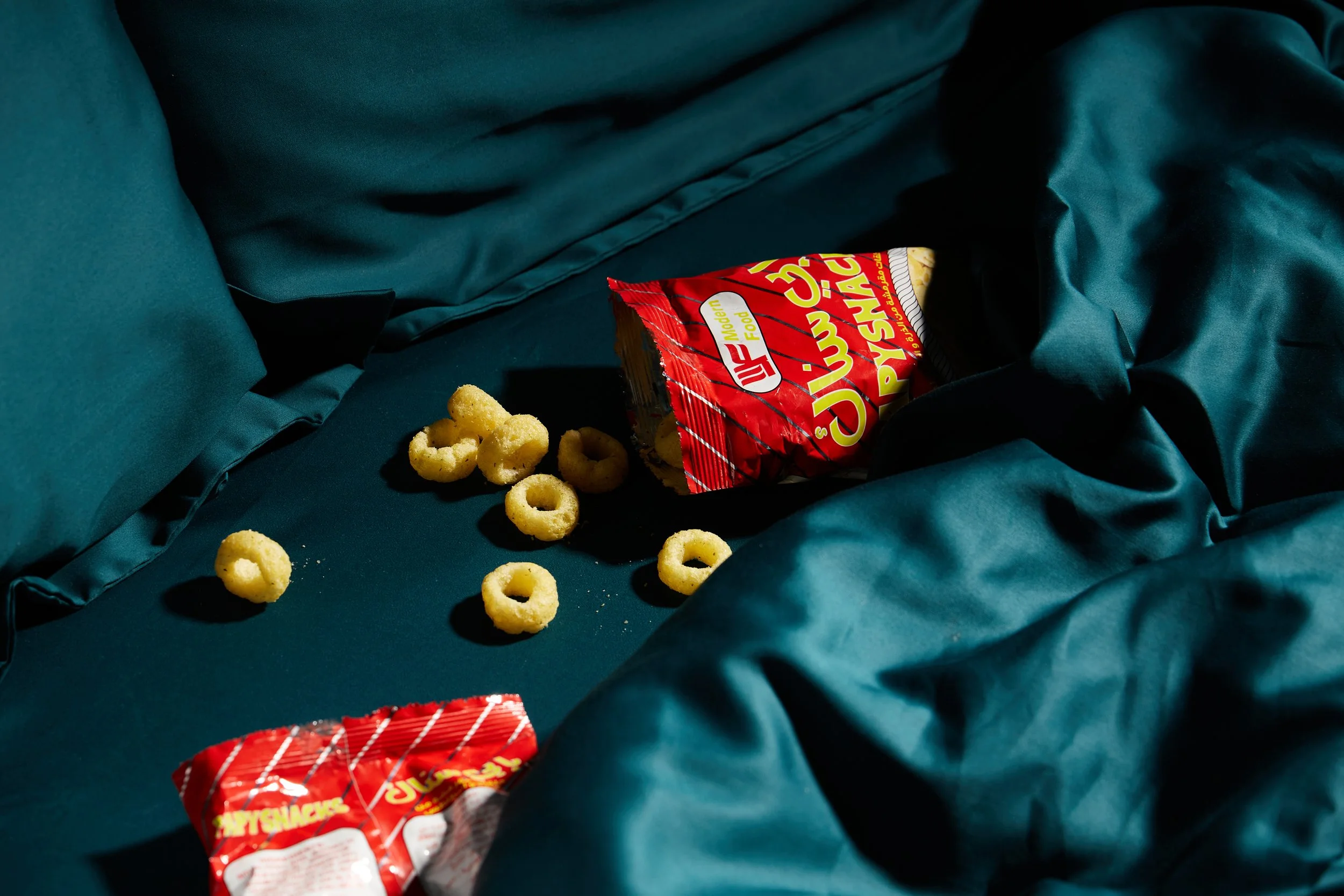 Open bag of Cheetos Puffs snack spilled on dark teal bed with some snack pieces scattered around.