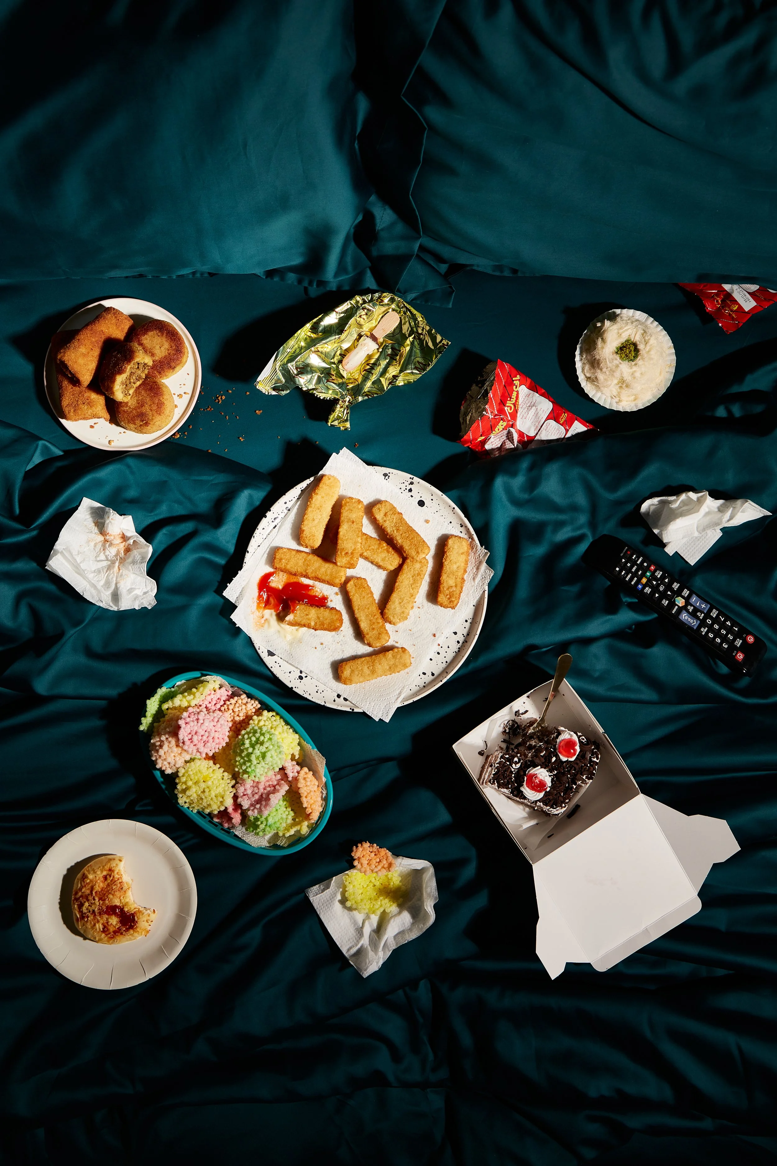 A messy display of various snacks and desserts on a dark green bedsheet, including fried snacks, a partially eaten pizza, canned fruit candies, a chocolate cake with cherries, and a pink dessert, along with a TV remote, a tissue, and a few crumpled n