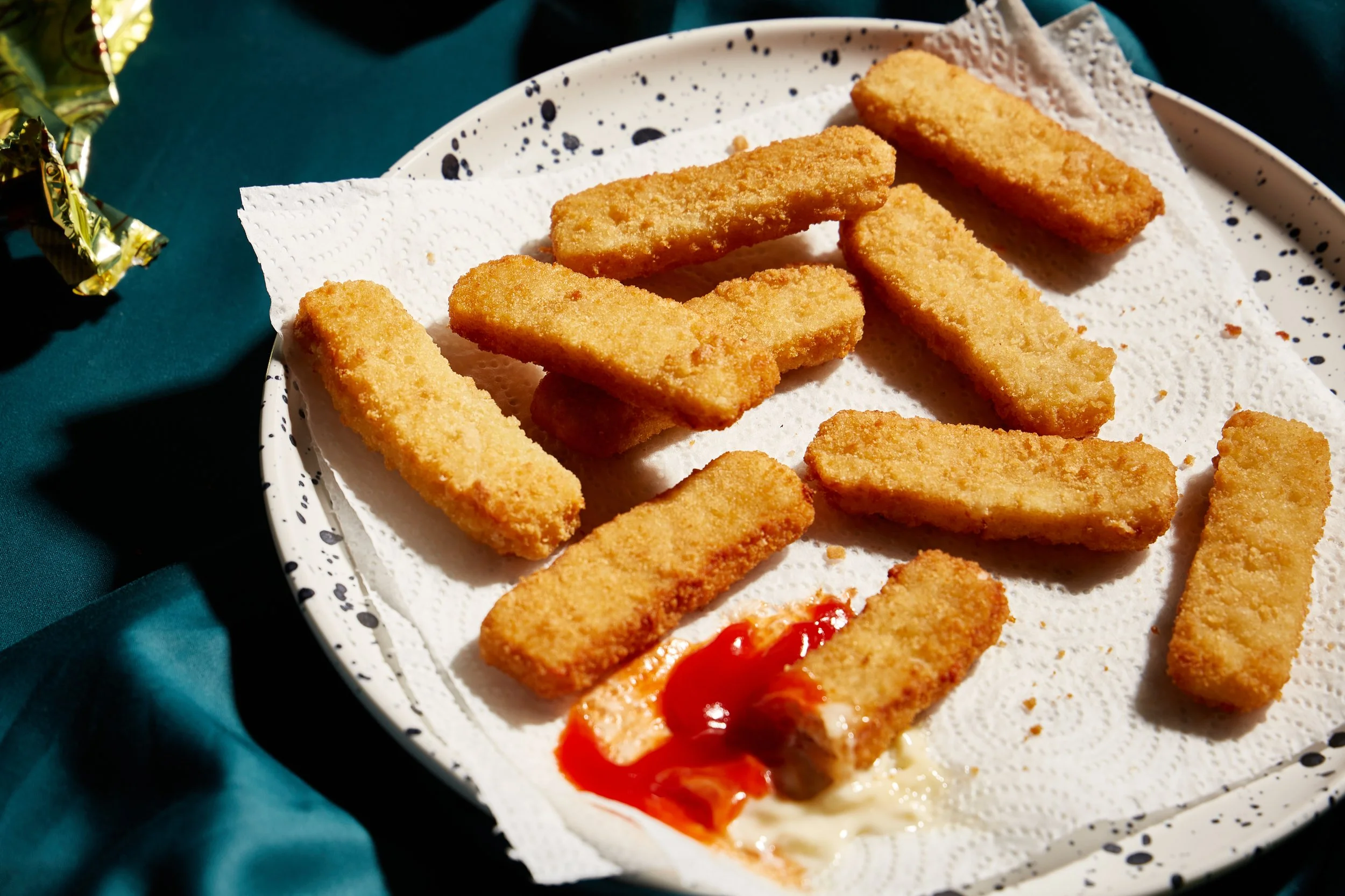 Plate of breaded chicken tenders with one partially eaten piece topped with ketchup and mayonnaise, on a paper towel-lined plate.