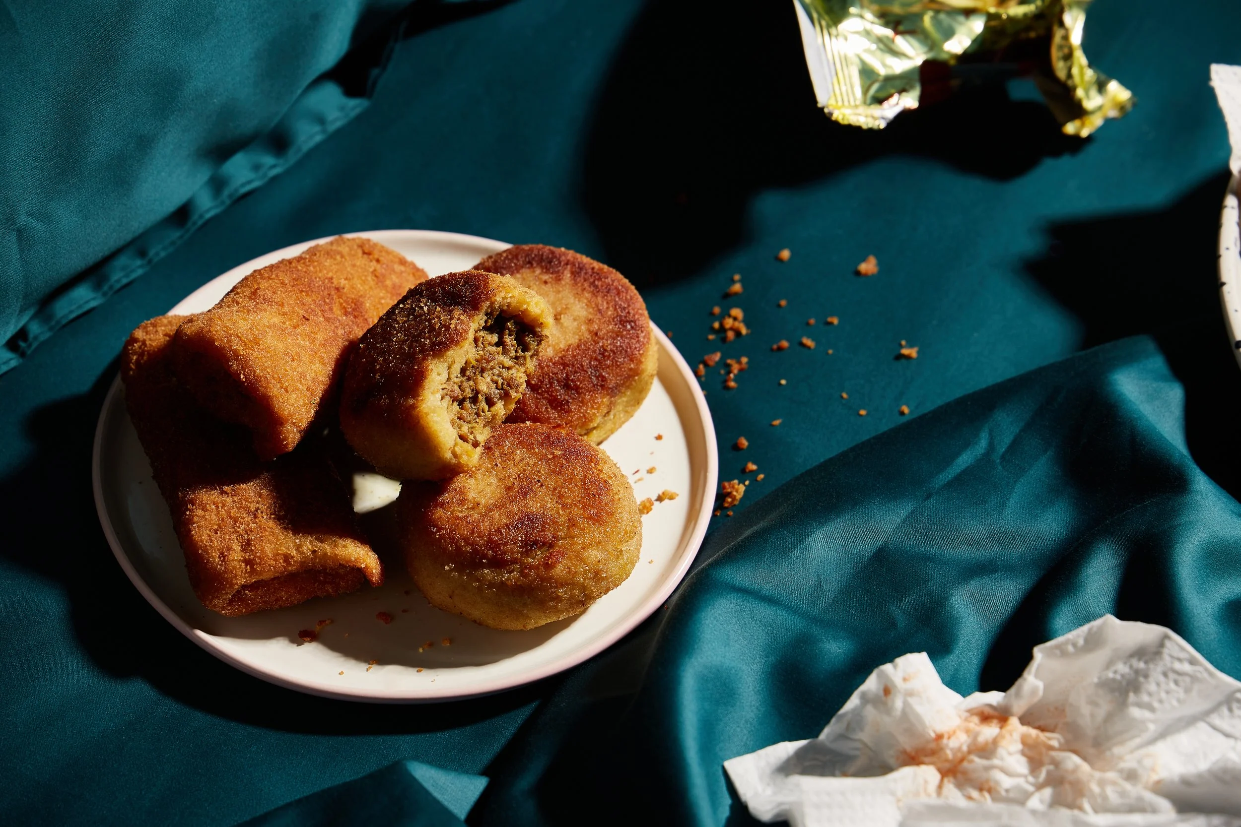 Fried meat-filled croquettes on a white plate, with one croquette cut open revealing the meat filling. The plate is on a dark teal fabric surface with scattered crumbs, and a crumpled paper napkin nearby.