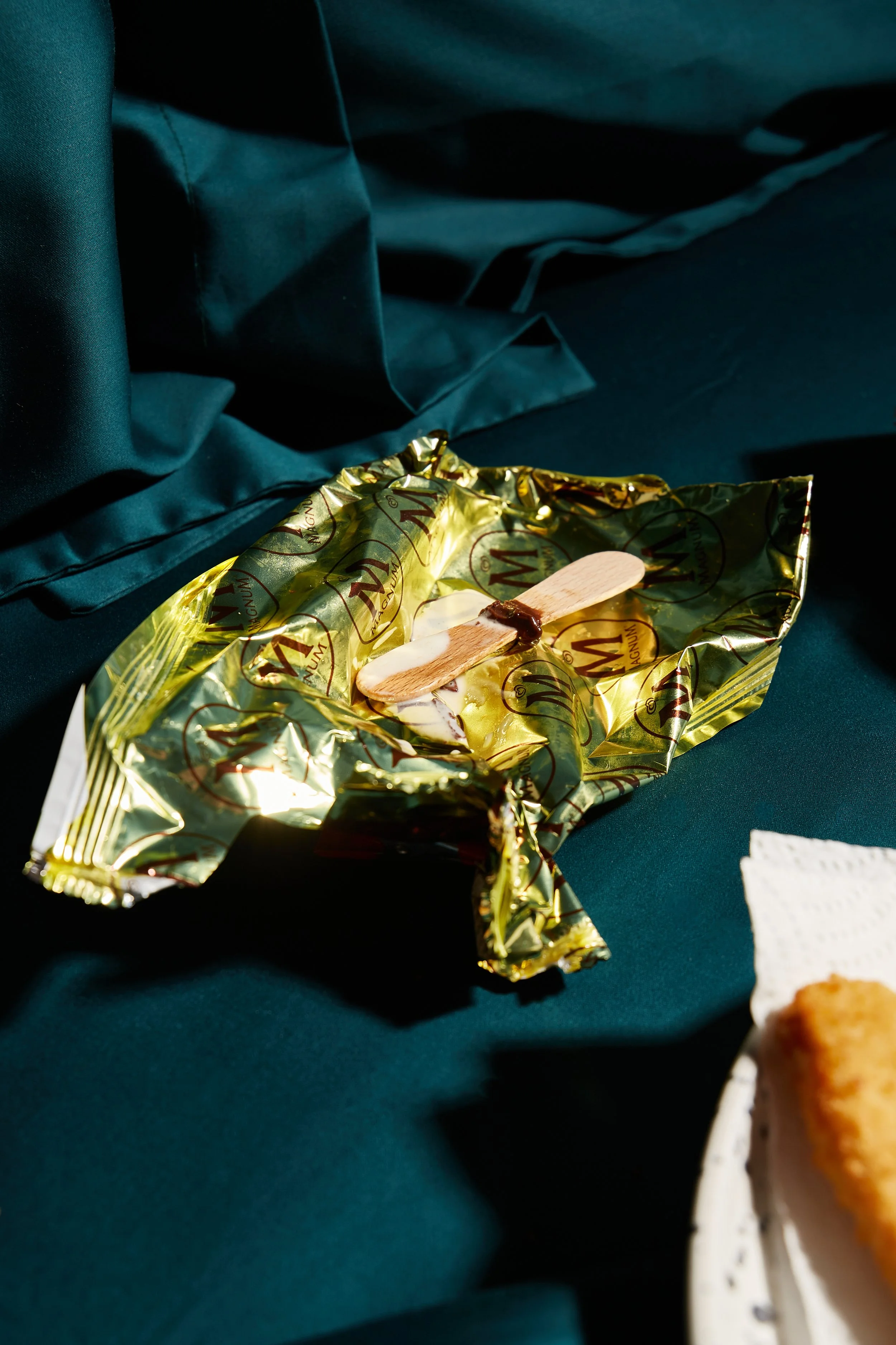 An unwrapped gold foil-wrapped ice cream bar with a wooden stick on a dark teal fabric surface, with part of a paper plate with fried food visible in the bottom right corner.