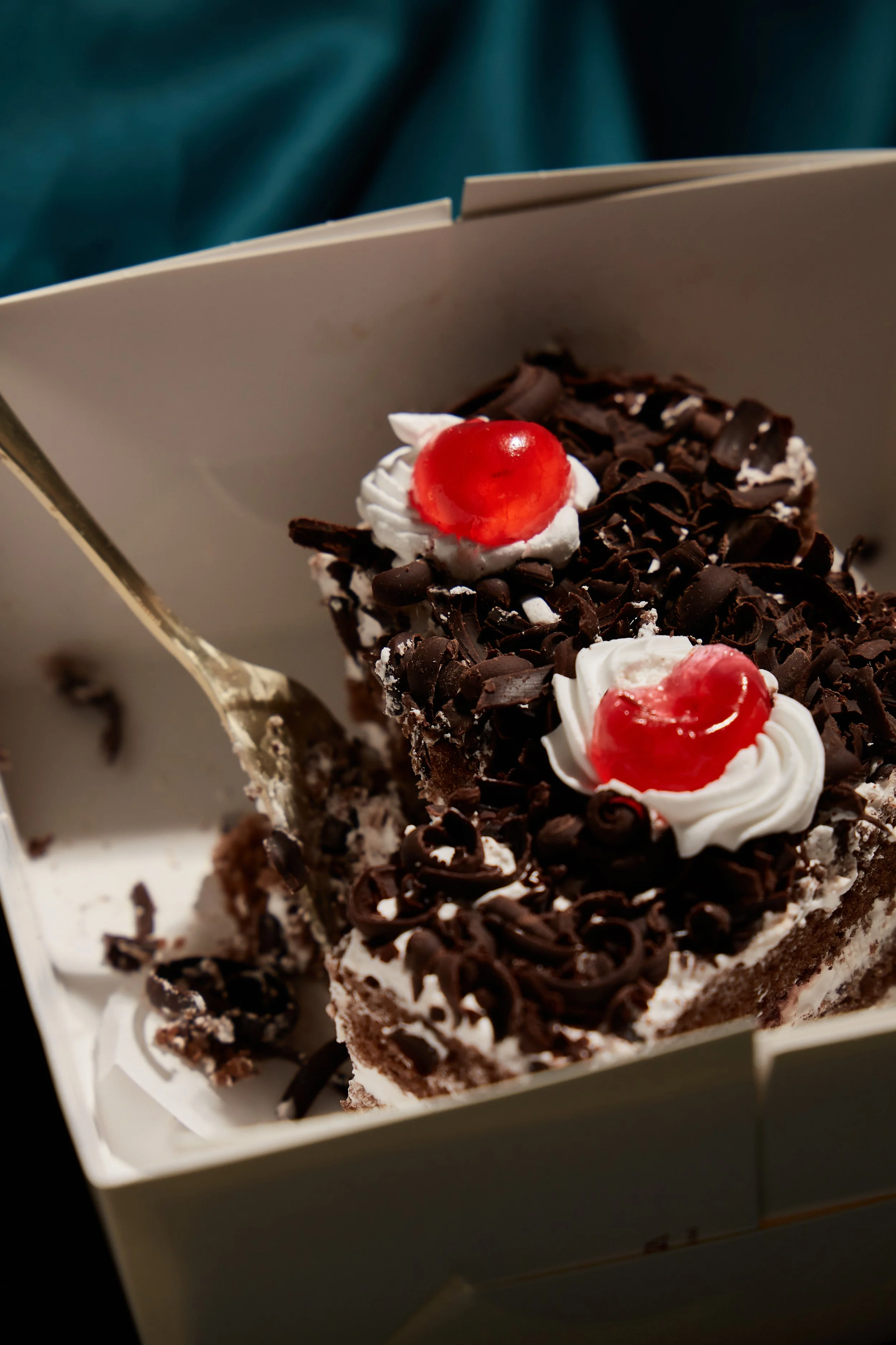 Slice of chocolate birthday cake decorated with whipped cream and cherries inside a white box.