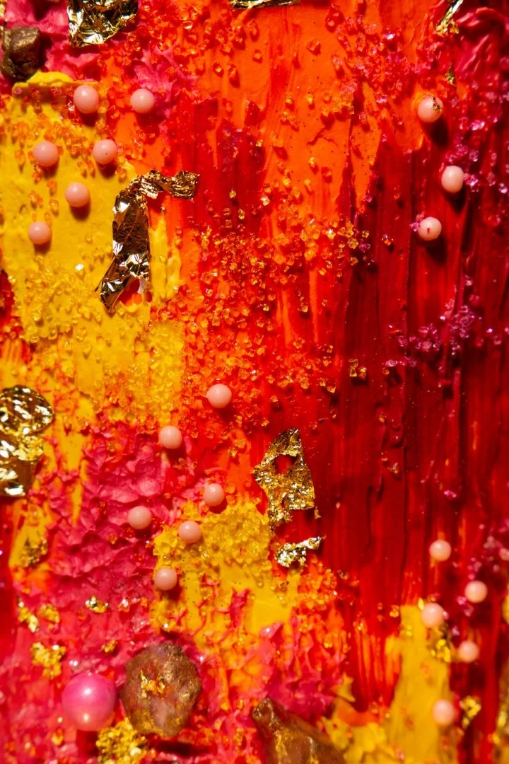 Close-up of colorful textured abstract artwork with red, orange, yellow, pink colors, gold foil, pink beads, and yellow and pink granules.