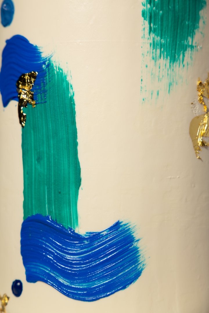 Abstract painting with broad vertical green, and blue brush strokes on a white wall, accented with gold foil details.