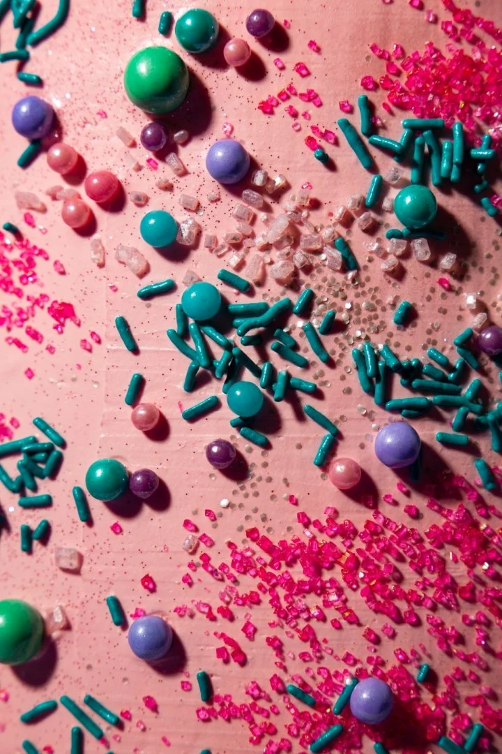 Close-up of colorful sprinkles and sugar crystals scattered on a pink surface, possibly on a cake or dessert.