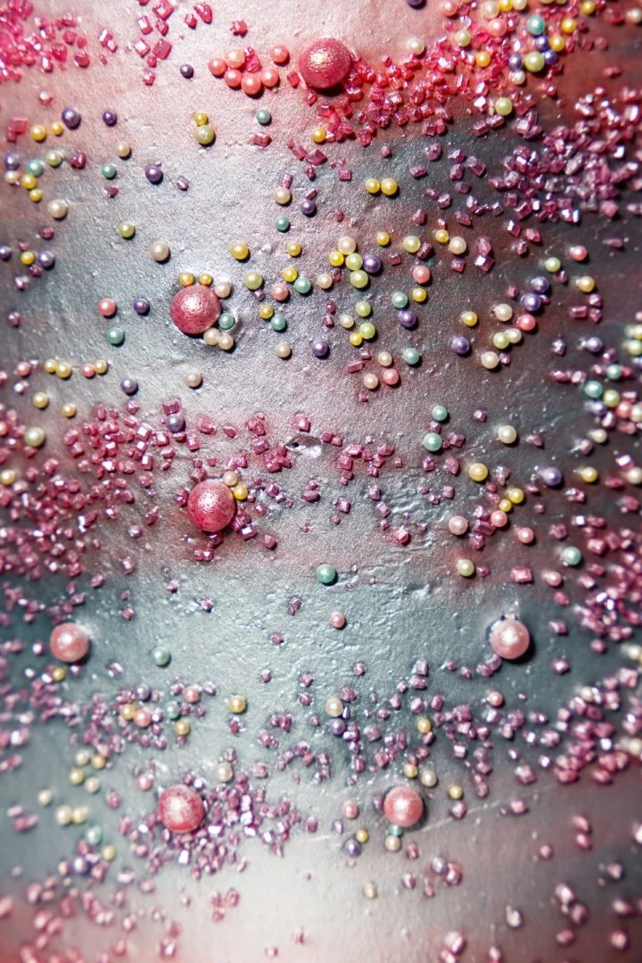 Close-up of colorful sprinkles and textured candies on a surface, featuring pink, purple, yellow, and green colors with metallic and matte finishes.