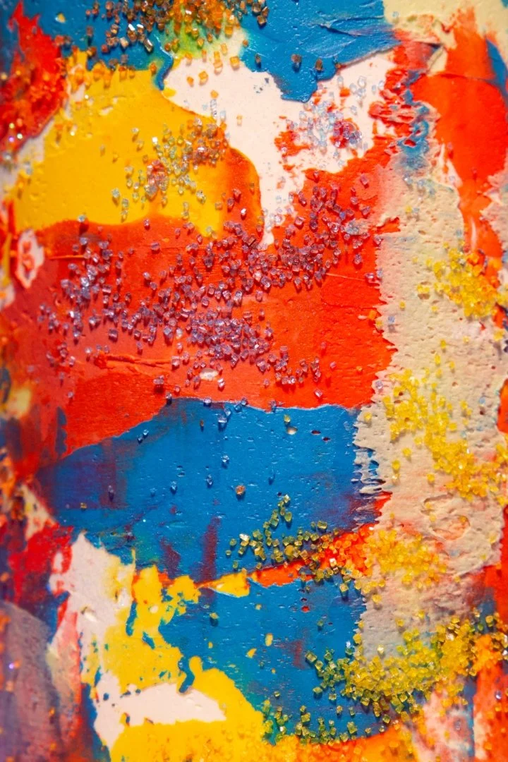 Close-up of colorful abstract painting with yellow, orange, red, blue, white, and green paint, with textured and sprinkled elements.
