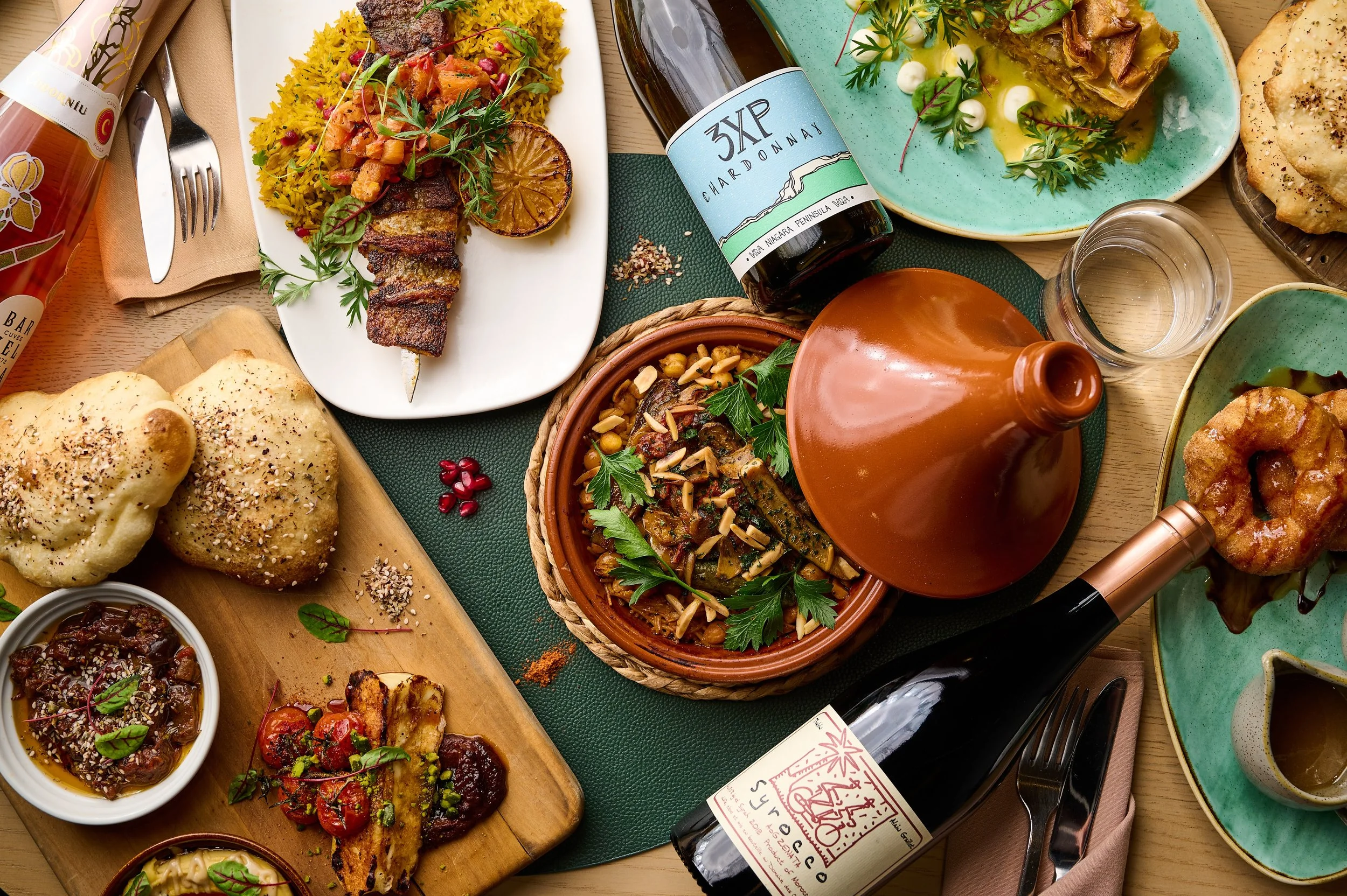 A table filled with assorted Middle Eastern dishes including grilled meat skewers, rice, dips, bread, baked goods, and wine bottles.