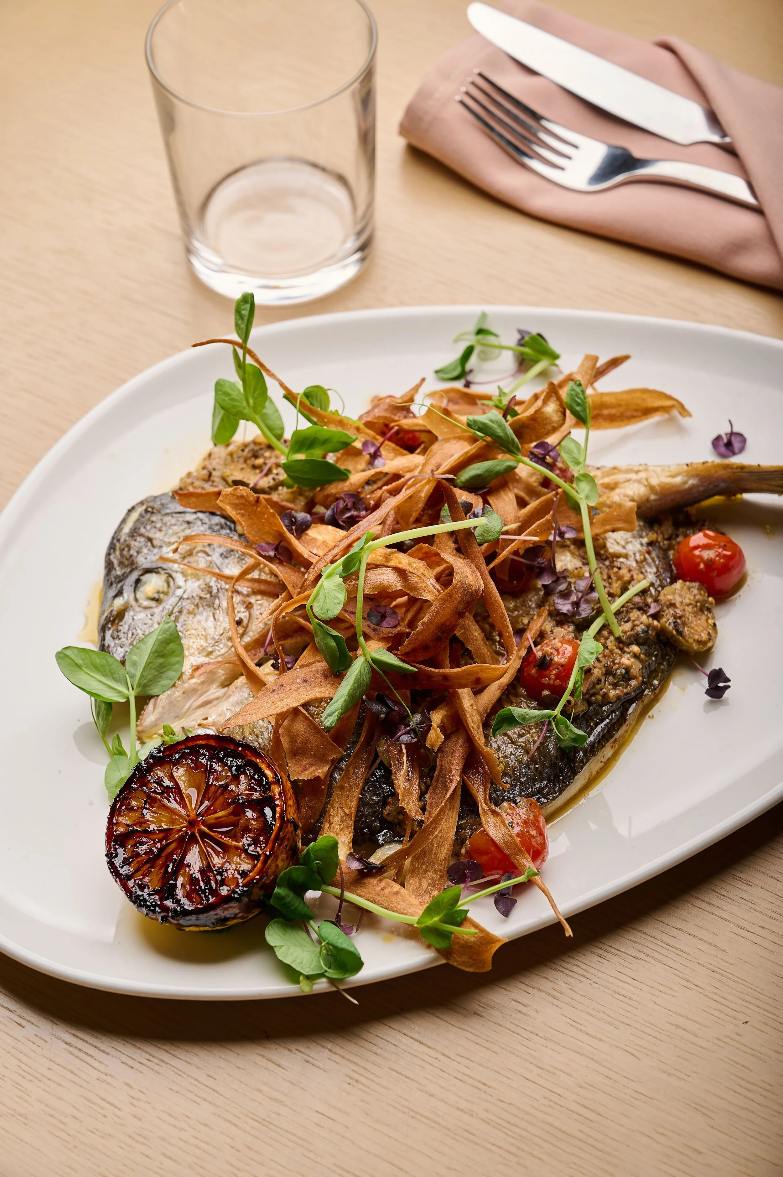 A plated grilled fish with garnishes, roasted lemon, cherry tomatoes, and herbs on a white dish, with a glass of water, fork, knife, and napkin on a wooden table.