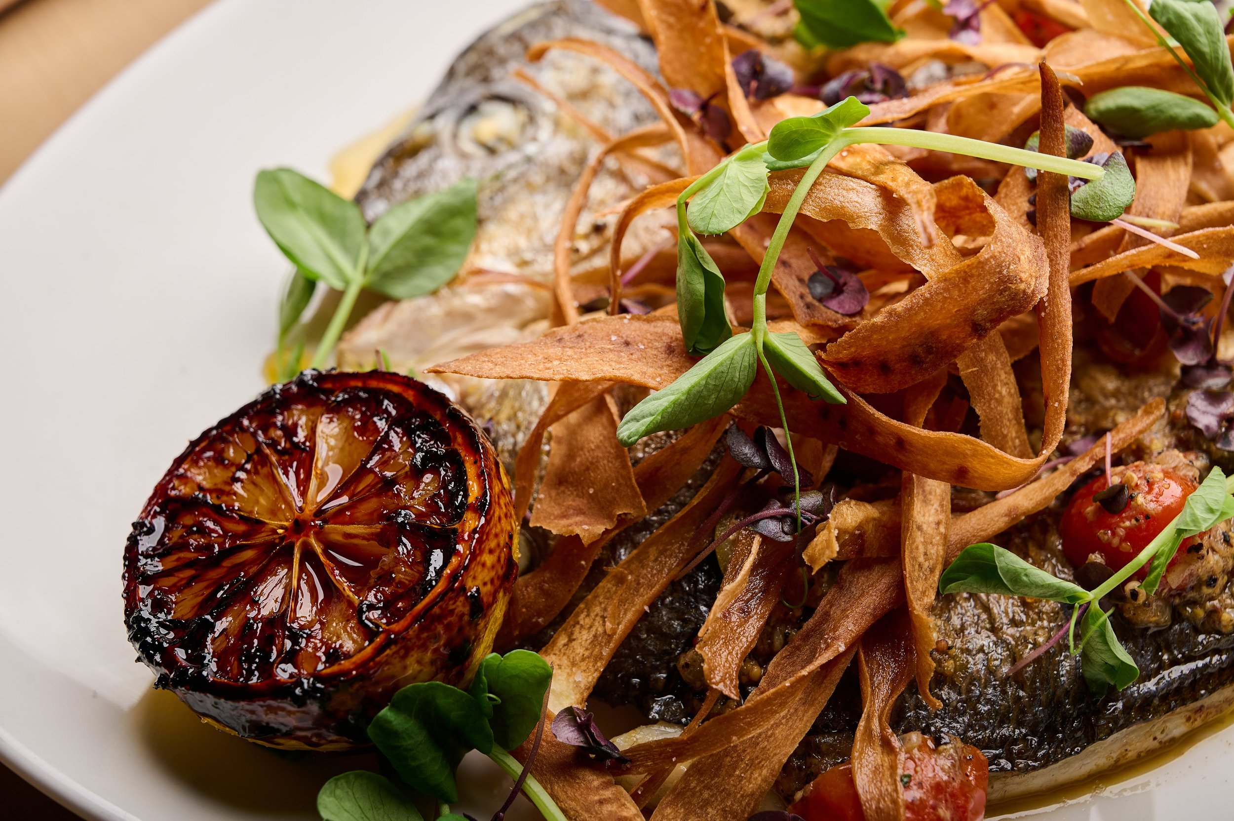 A plate of grilled fish topped with crispy fried onion strings, cherry tomatoes, grilled lemon half, and garnished with microgreens.