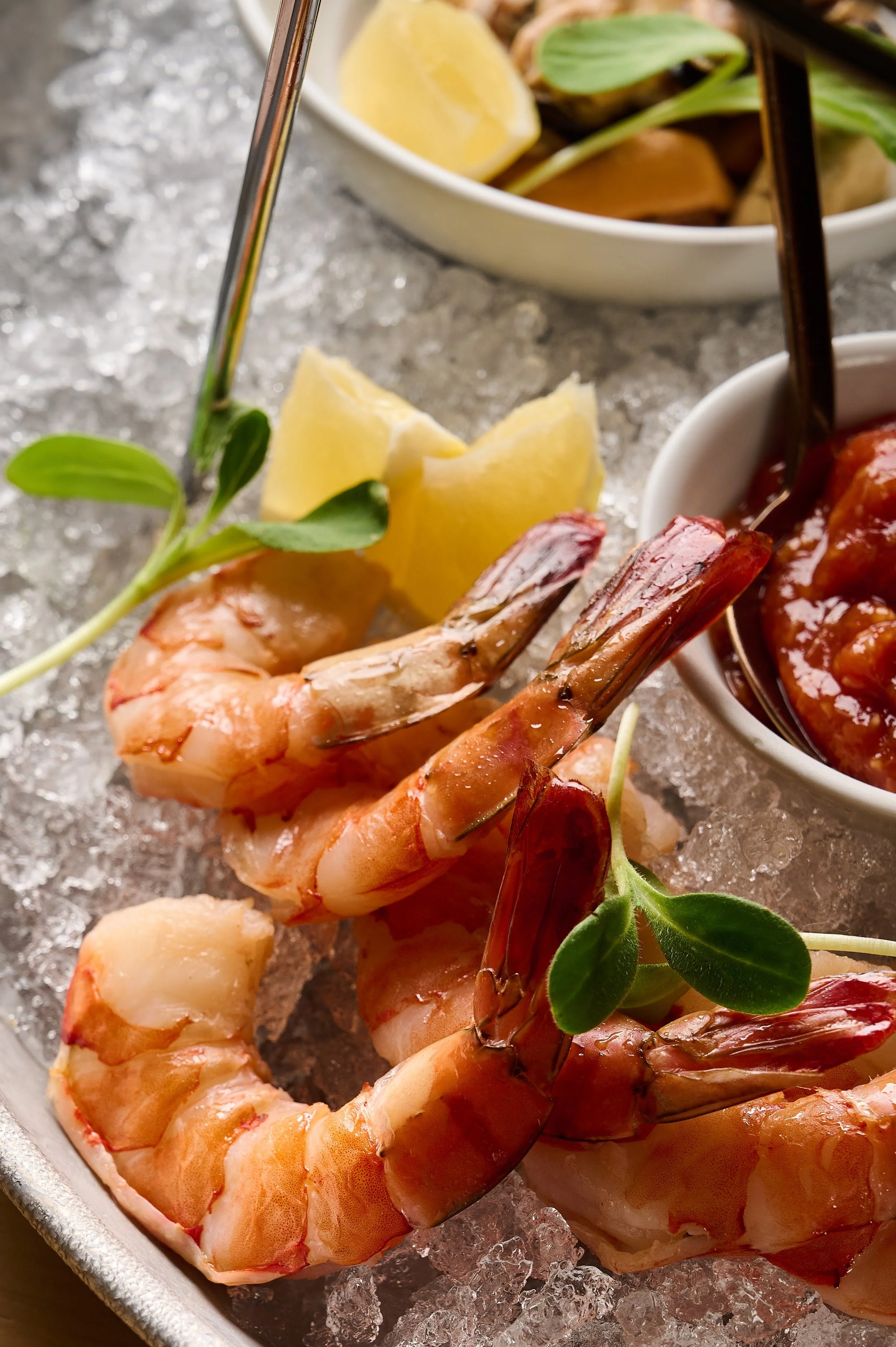 Cooked shrimp garnished with green leaves, lemon wedges, and served with dipping sauces on ice.