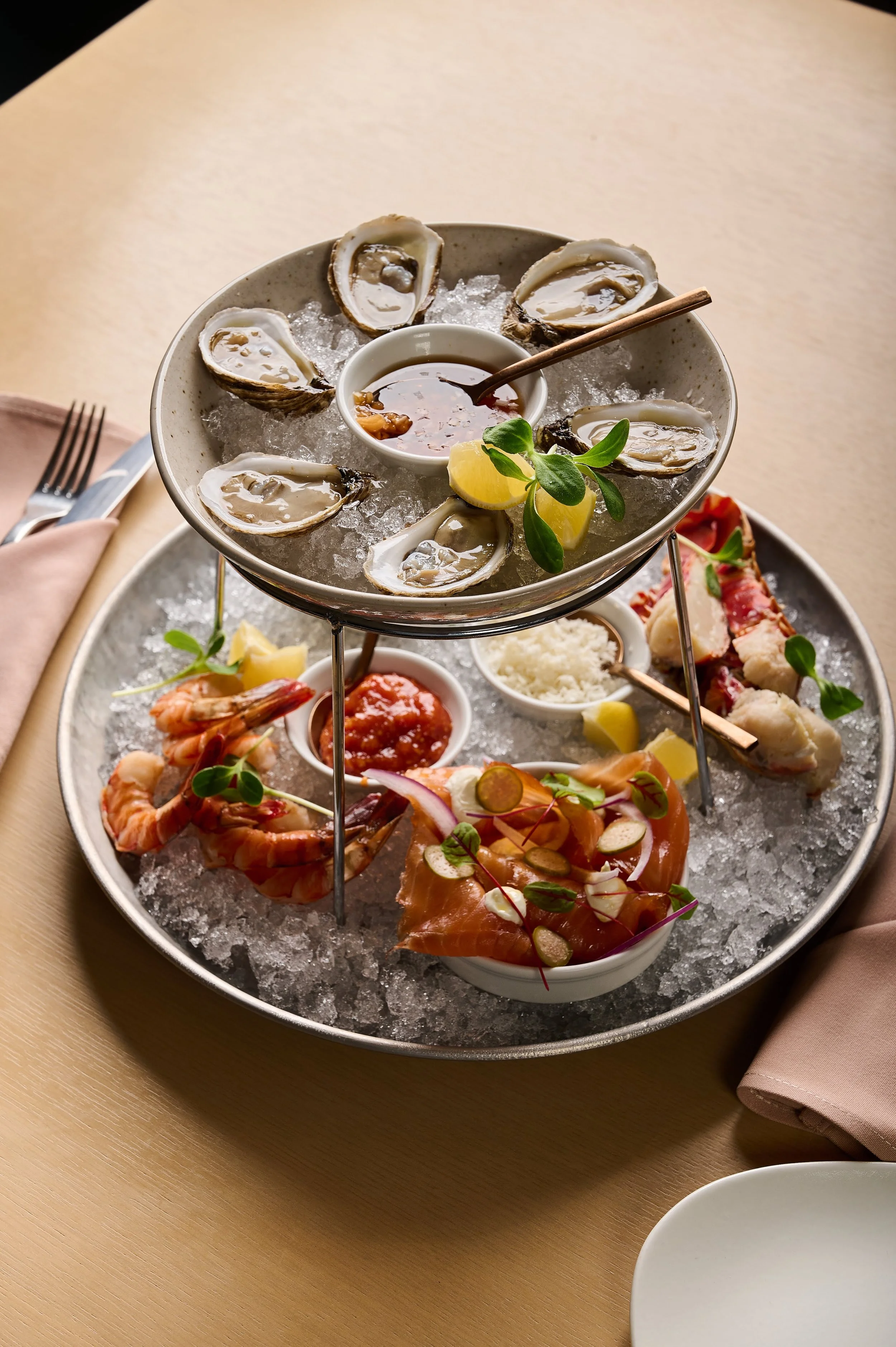 A seafood platter with oysters on the top tier, shrimp, and slices of cured meat on the bottom tier, served on a bed of ice with lemon wedges and garnished with herbs.