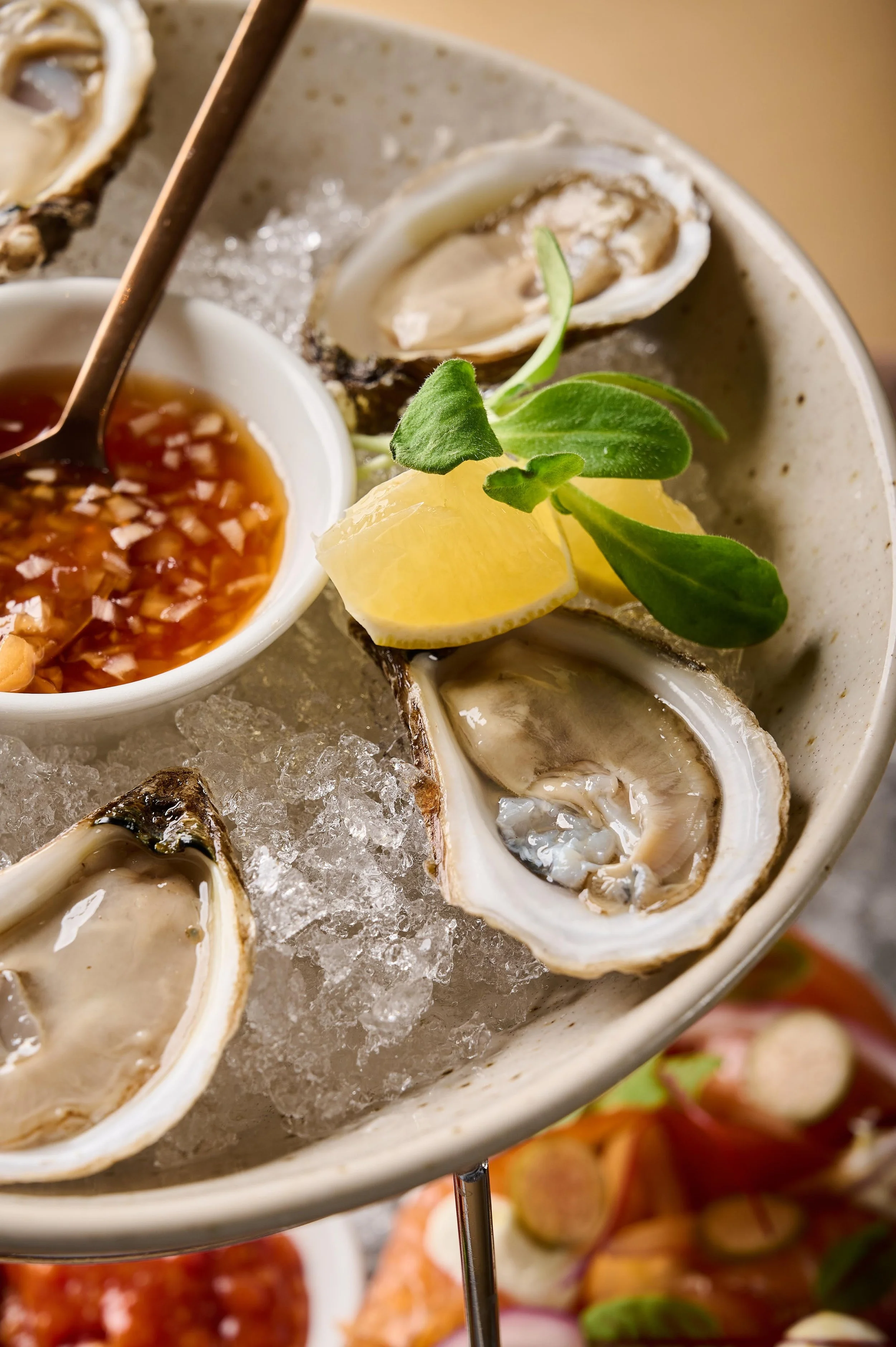 A platter of fresh oysters on ice with lemon wedges, a dipping sauce, and garnish.