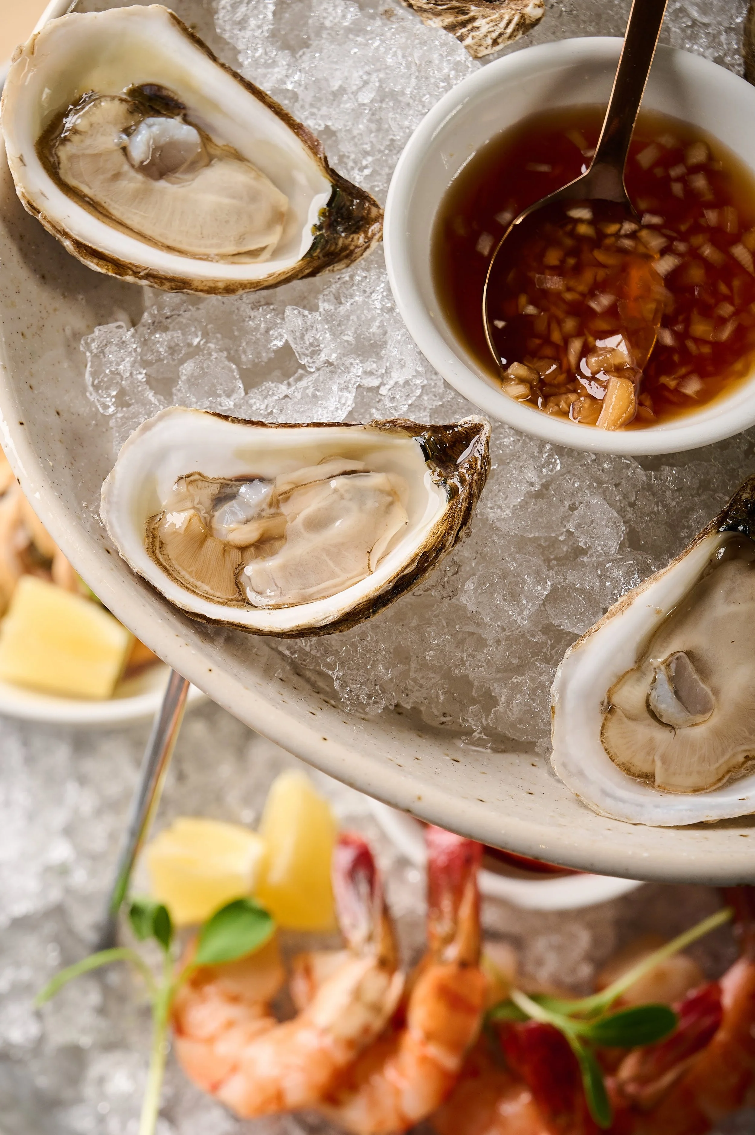 Fresh oysters on ice with a small bowl of mignonette sauce and lemon wedges, with shrimp and garnishes in the background.