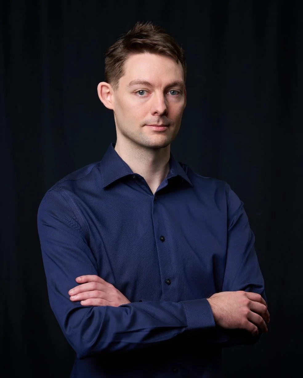 A young man with short brown hair, fair skin, wearing a dark blue button-up shirt, standing with arms crossed against a black background.