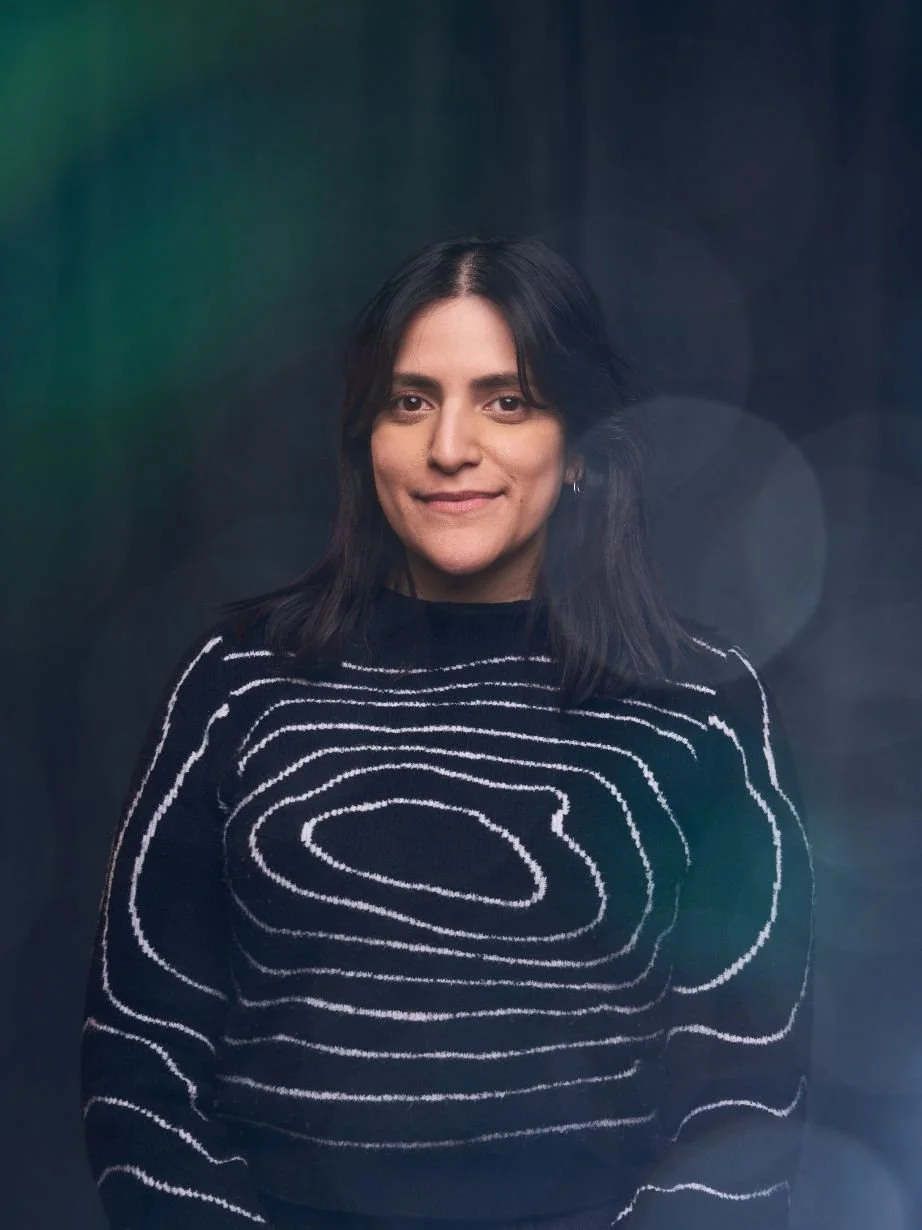 A woman with shoulder-length dark hair wearing a black sweater with white abstract line patterns, standing against a dark background with colorful bokeh lights.