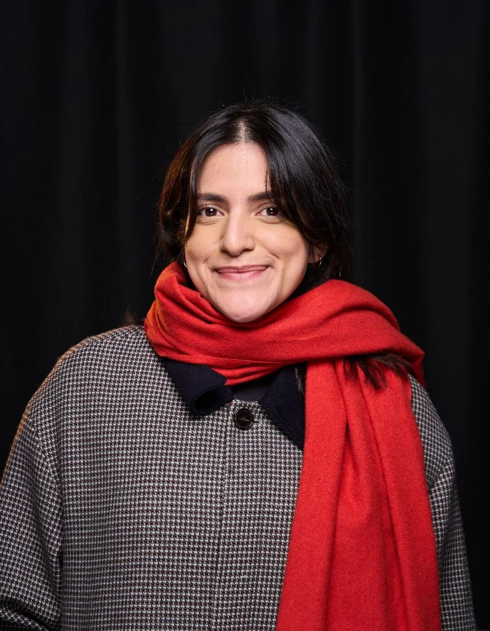 A woman with shoulder-length dark hair, wearing a checkered jacket and a red scarf, smiling in front of a black background.