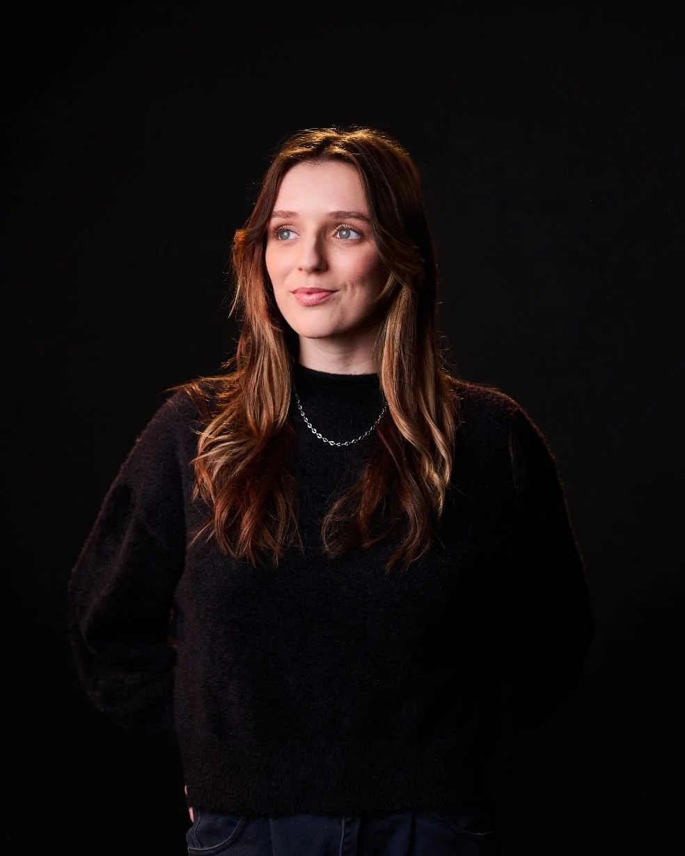 Portrait of a young woman with long, wavy brown hair and blue eyes, wearing a black sweater and a silver necklace, standing against a black background.