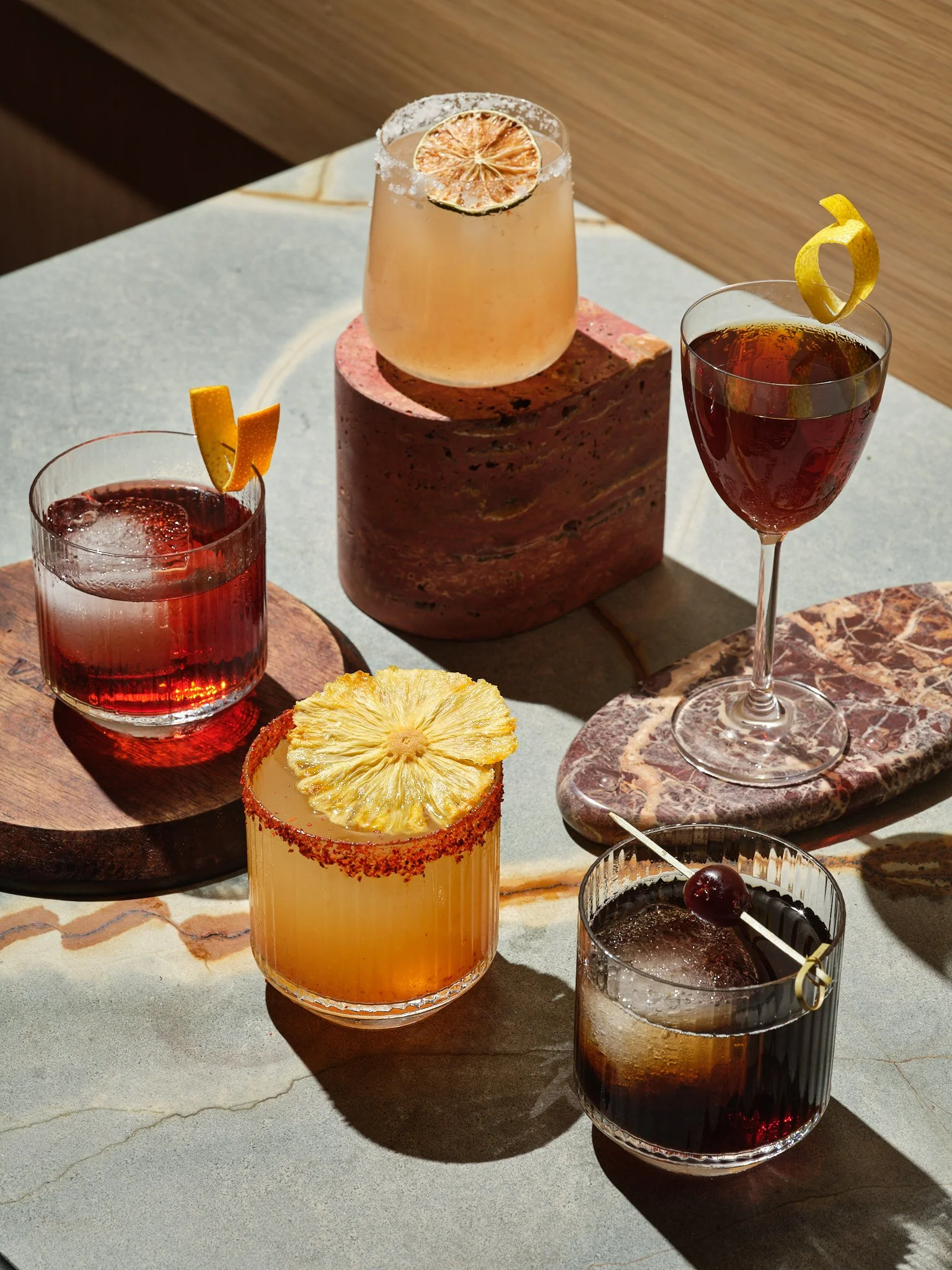 Four colorful cocktails with garnishes, arranged on different surfaces on a light-colored stone table.