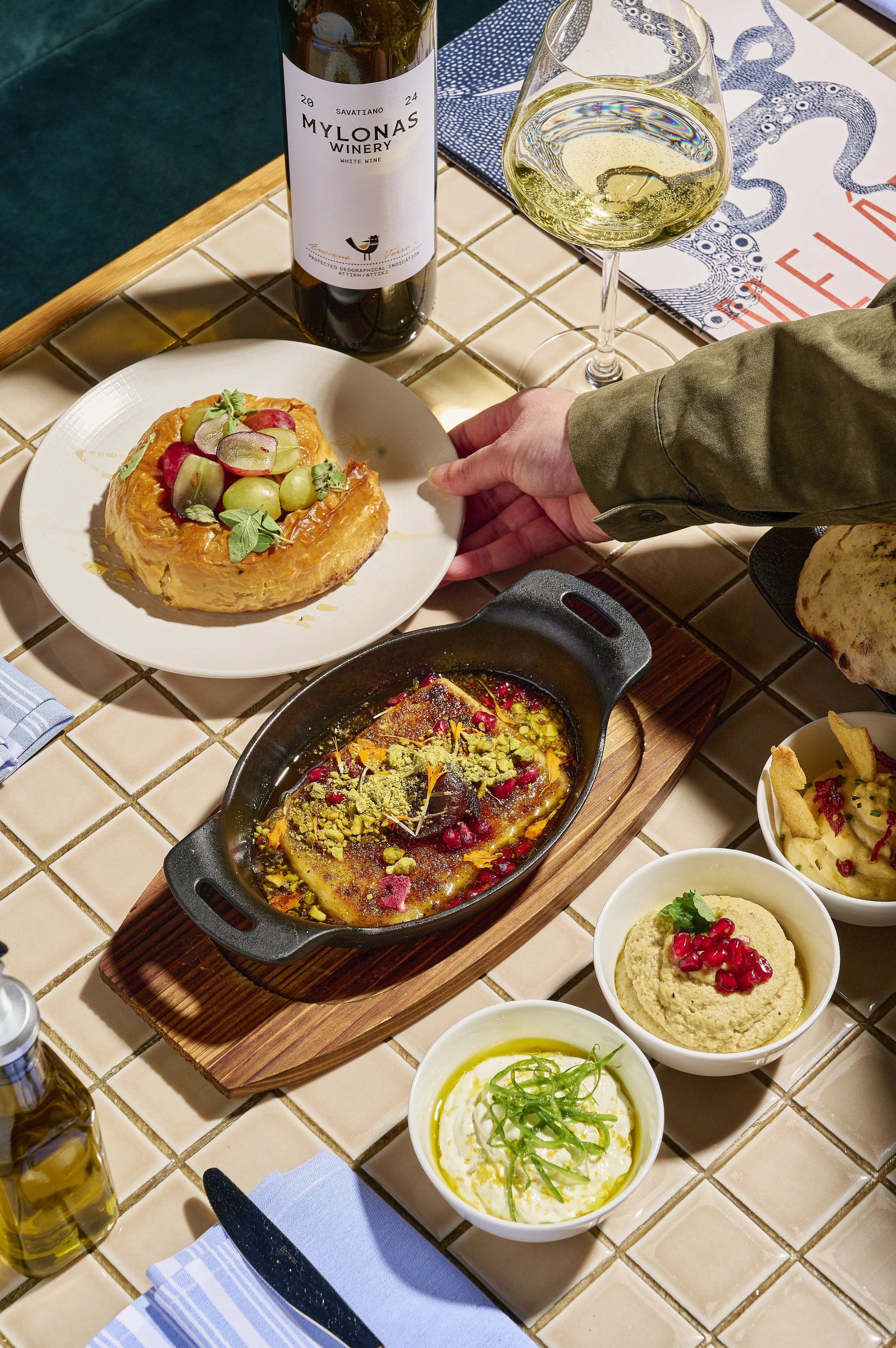 A table with various dishes including a fruit tart topped with grapes and radishes, a cast iron skillet with cooked fish garnished with pomegranate seeds and herbs, bowls of hummus and other dips, a glass of white wine, a bottle of wine, and place se