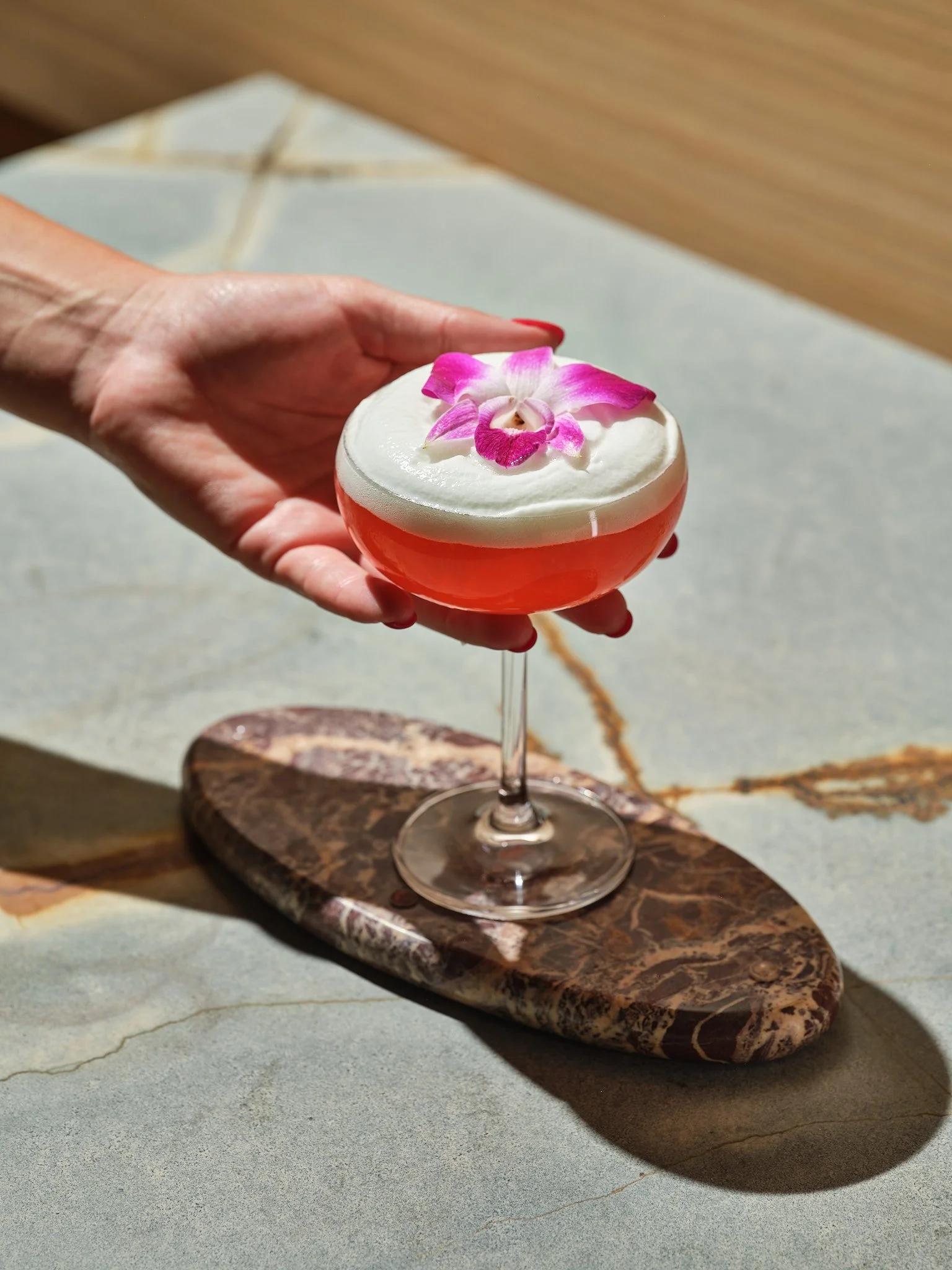 A hand holding a cocktail glass with a pink drink, topped with white foam and a pink orchid flower, placed on a marble serving board.