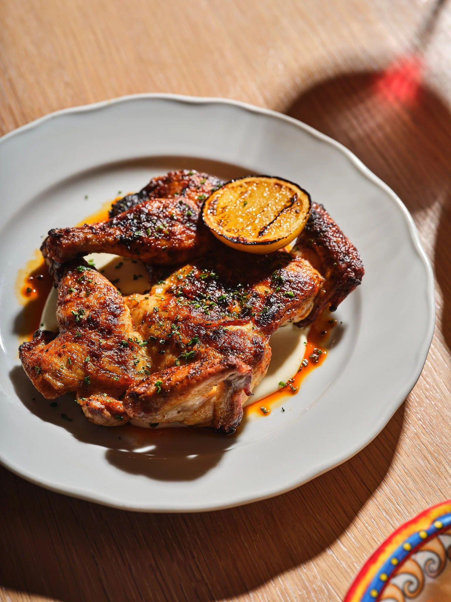 Grilled chicken thigh on a white plate with charred lemon half, chopped herbs, and sauce, set on a wooden table.