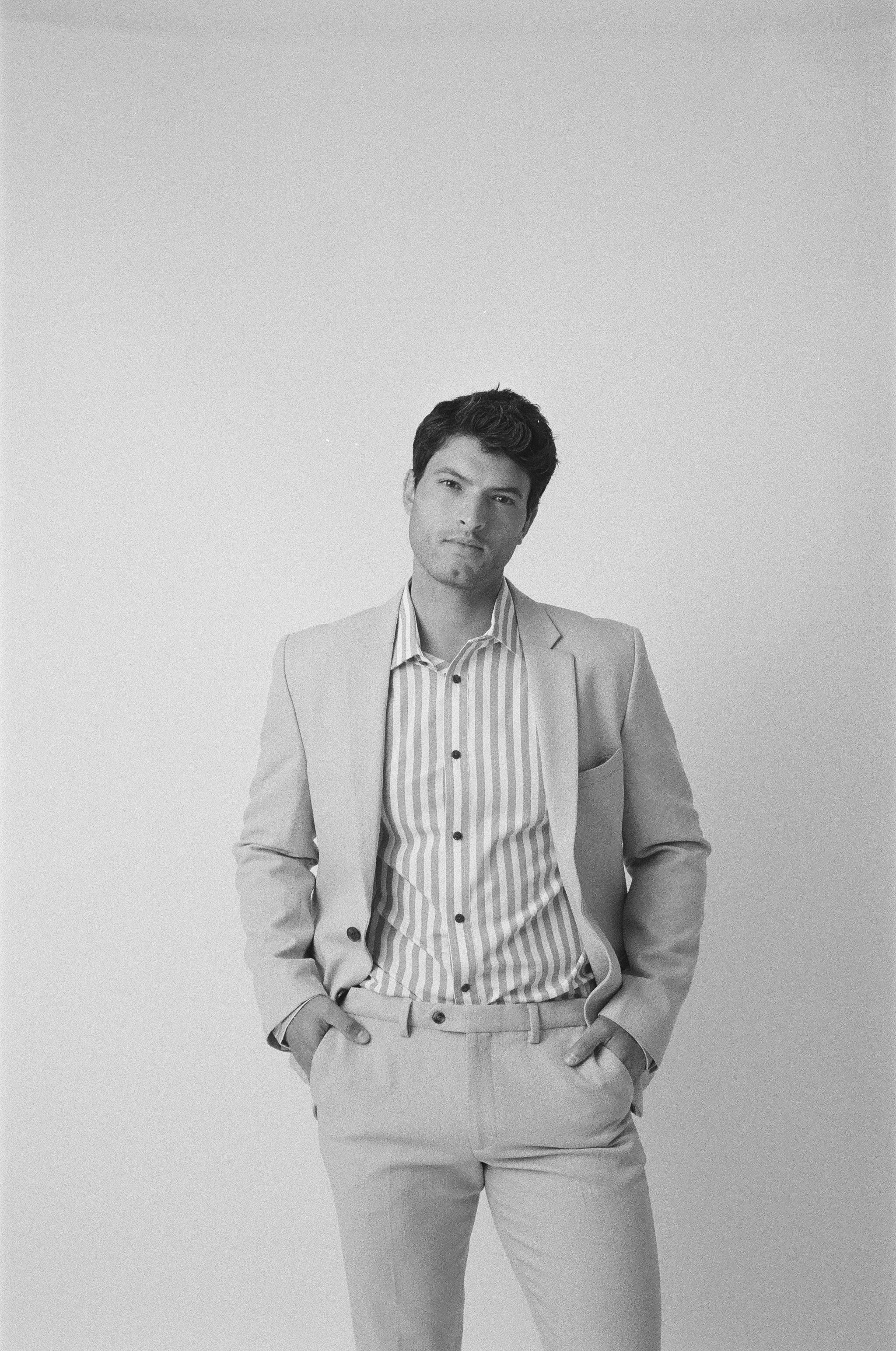A young man in a light suit with hands in pockets, wearing a striped shirt, standing against a plain light background, looking confidently at the camera.
