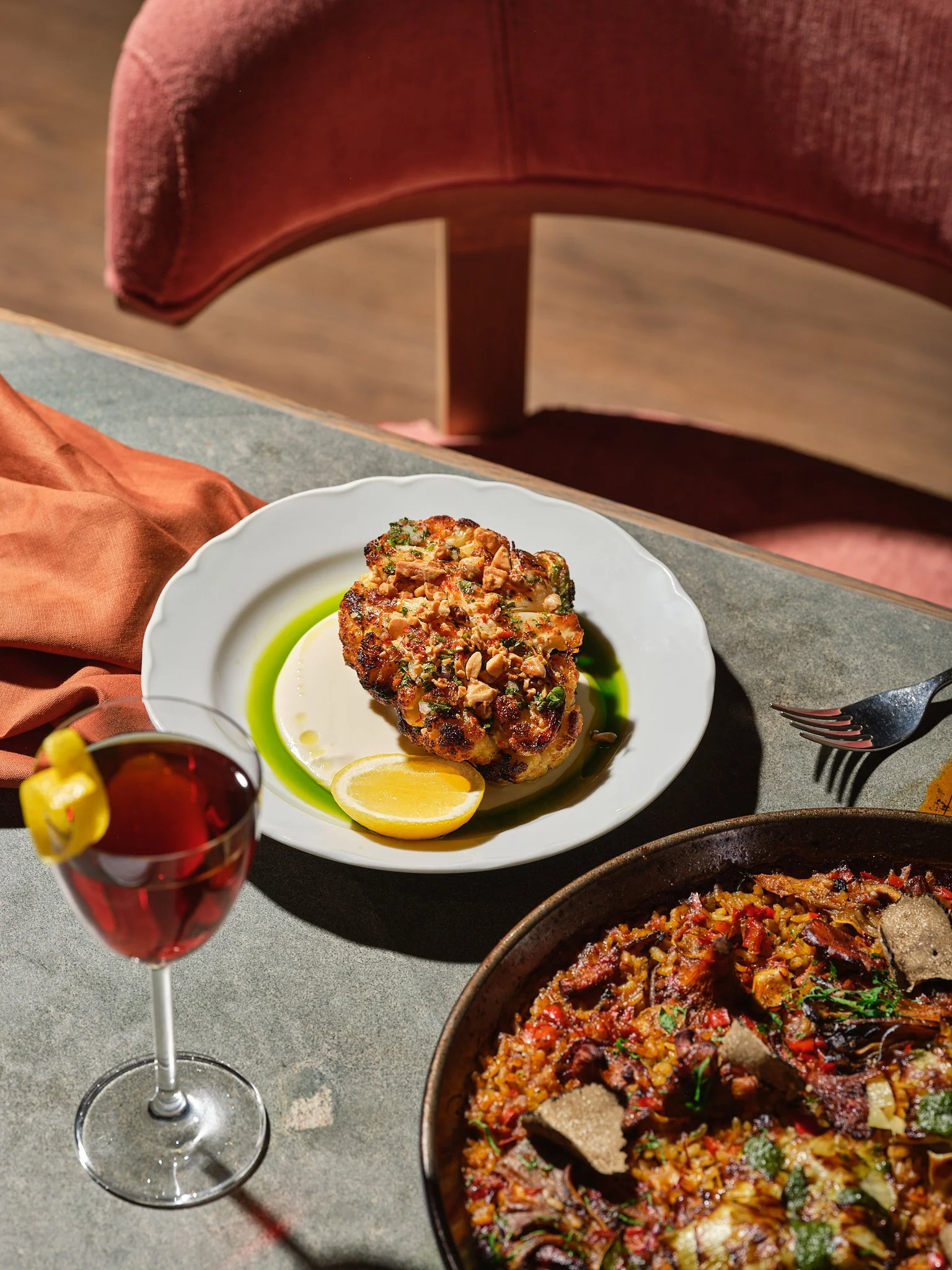 A plate of roasted cauliflower topped with chopped nuts and herbs, garnished with a lemon half, on a table with a slice of lemon, a glass of red drink, and a skillet of cooked rice and vegetables.