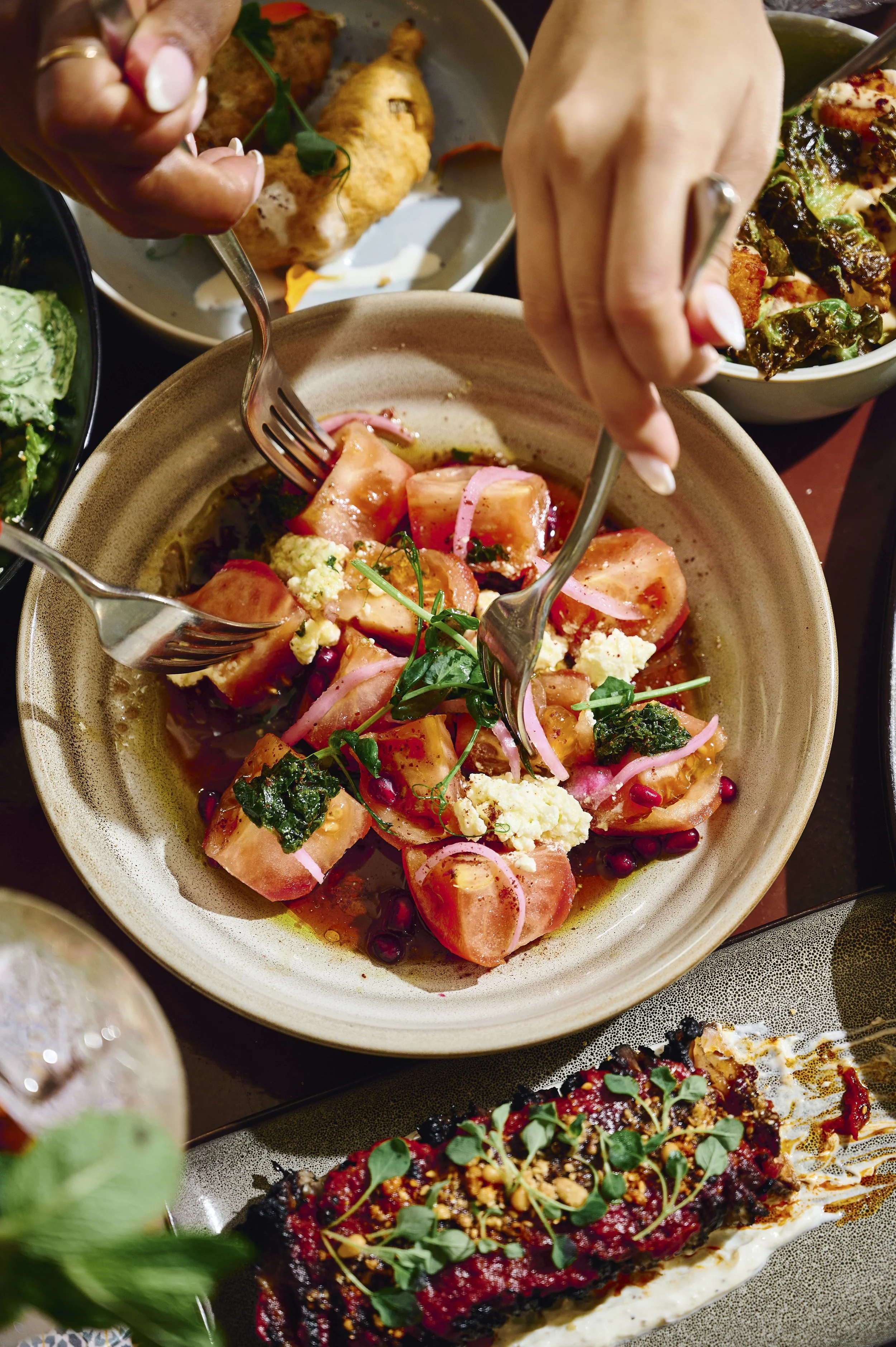 Close-up of a tomato salad with pink onions, herbs, and crumbled cheese in a beige bowl, surrounded by other dishes on a table.