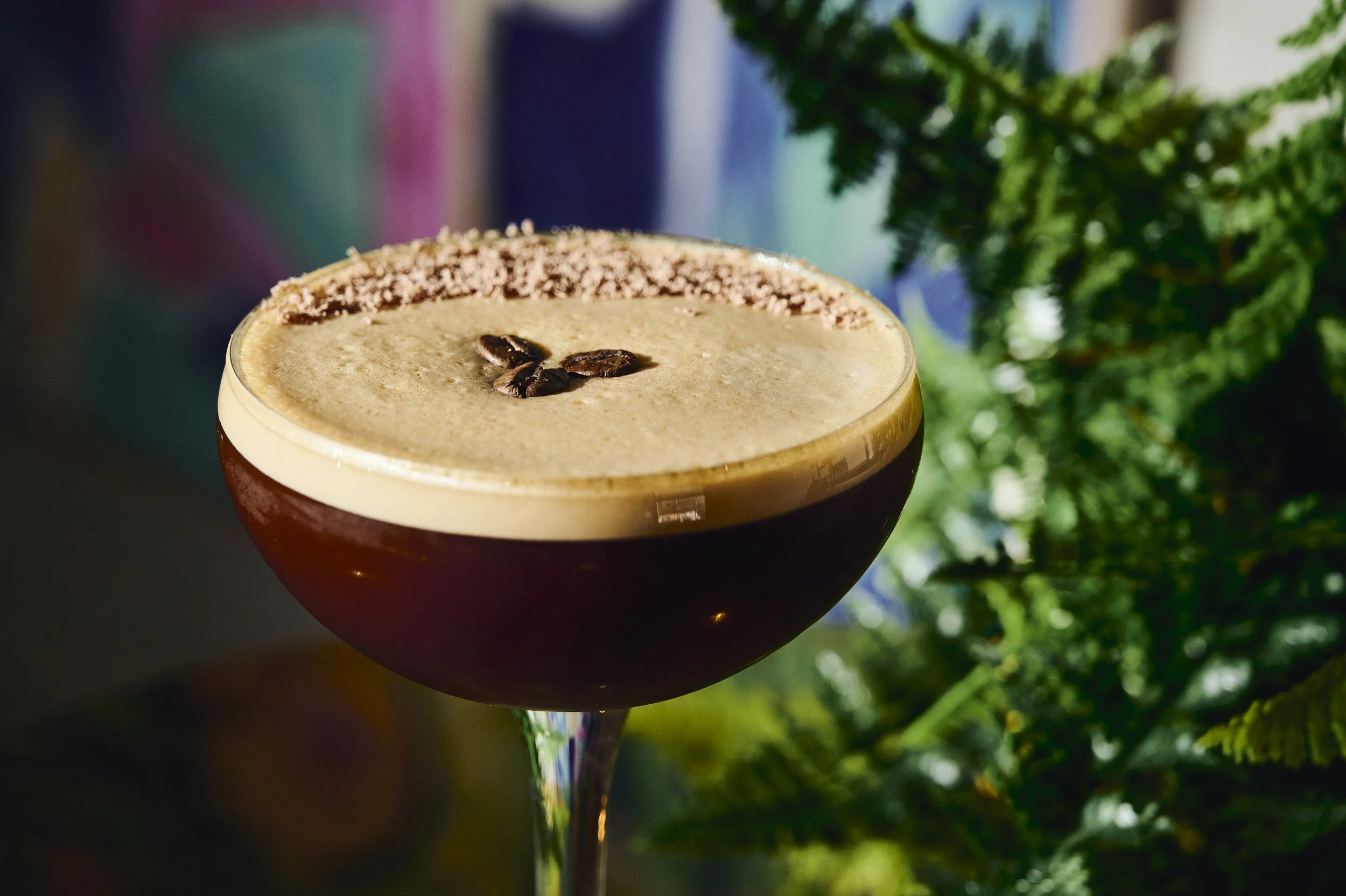 Close-up of a dark coffee cocktail with foam and coffee beans on top, garnished with a pink rim, against a background of green foliage and colorful blurred objects.