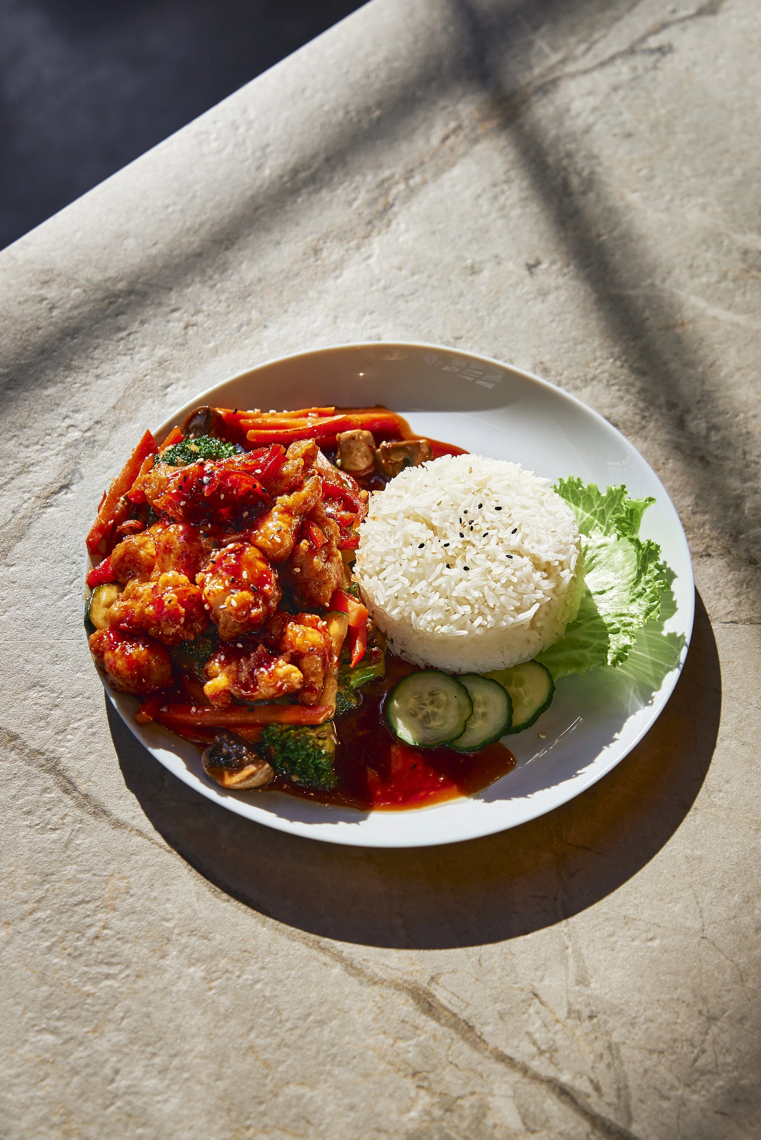 Plate of Asian cuisine with rice, stir-fry vegetables, and chicken in a savory sauce on a white plate, garnished with cucumber slices and lettuce.