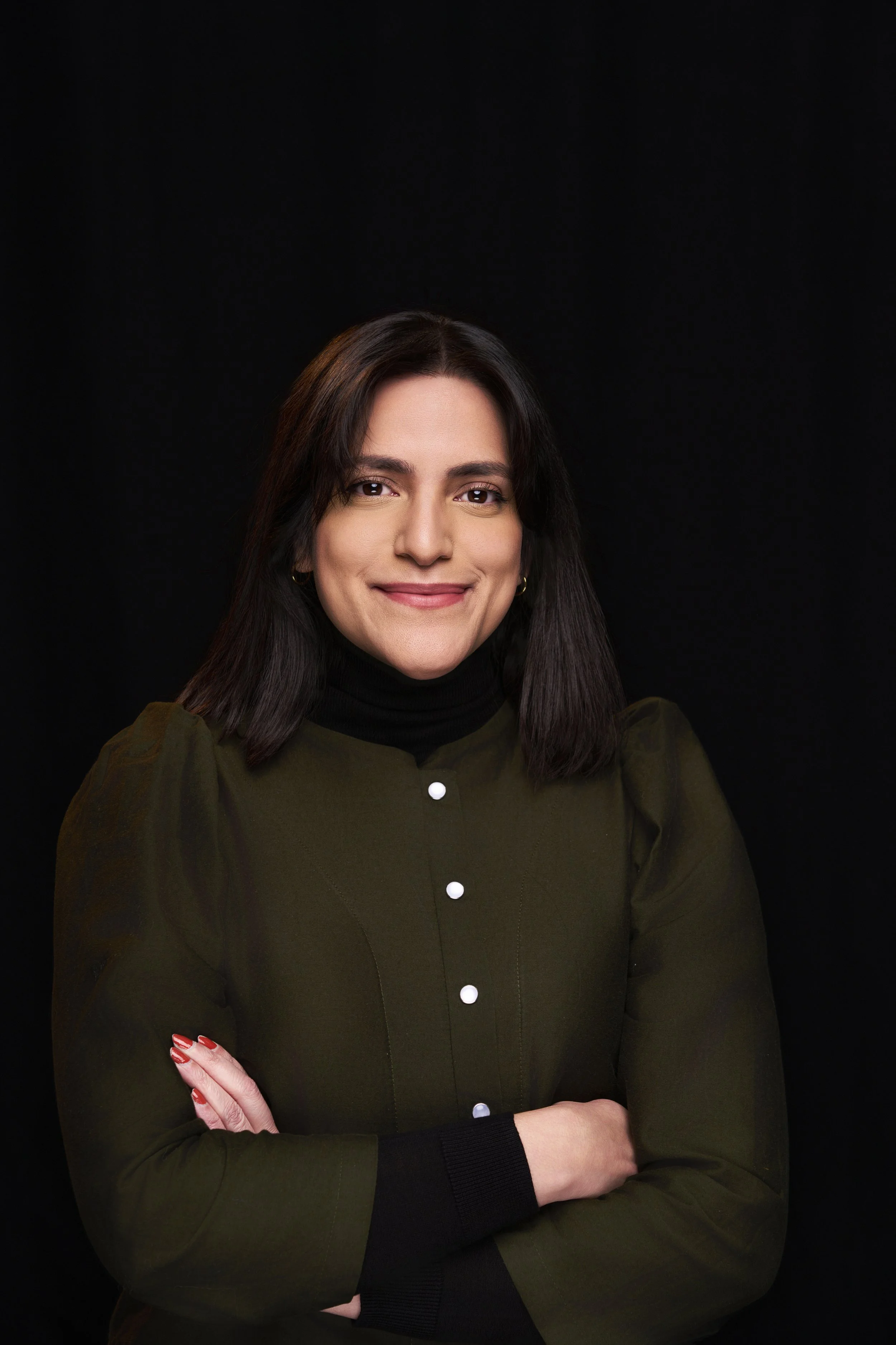 A woman with dark brown hair smiling, wearing a dark green button-up shirt with a black turtleneck underneath, standing against a black background.