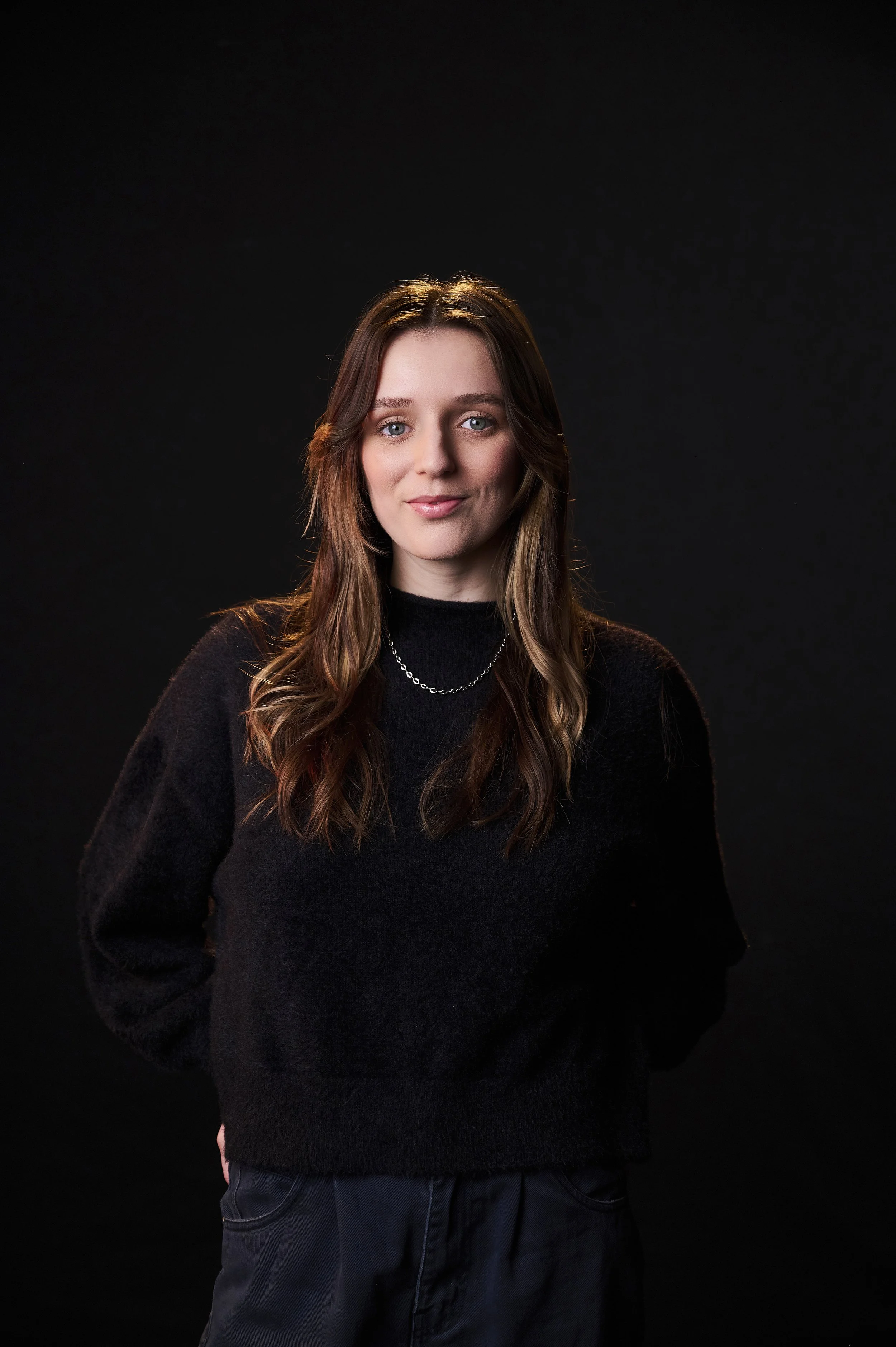 A young woman with long wavy brown hair, wearing a black sweater and a silver chain necklace, stands against a dark background.