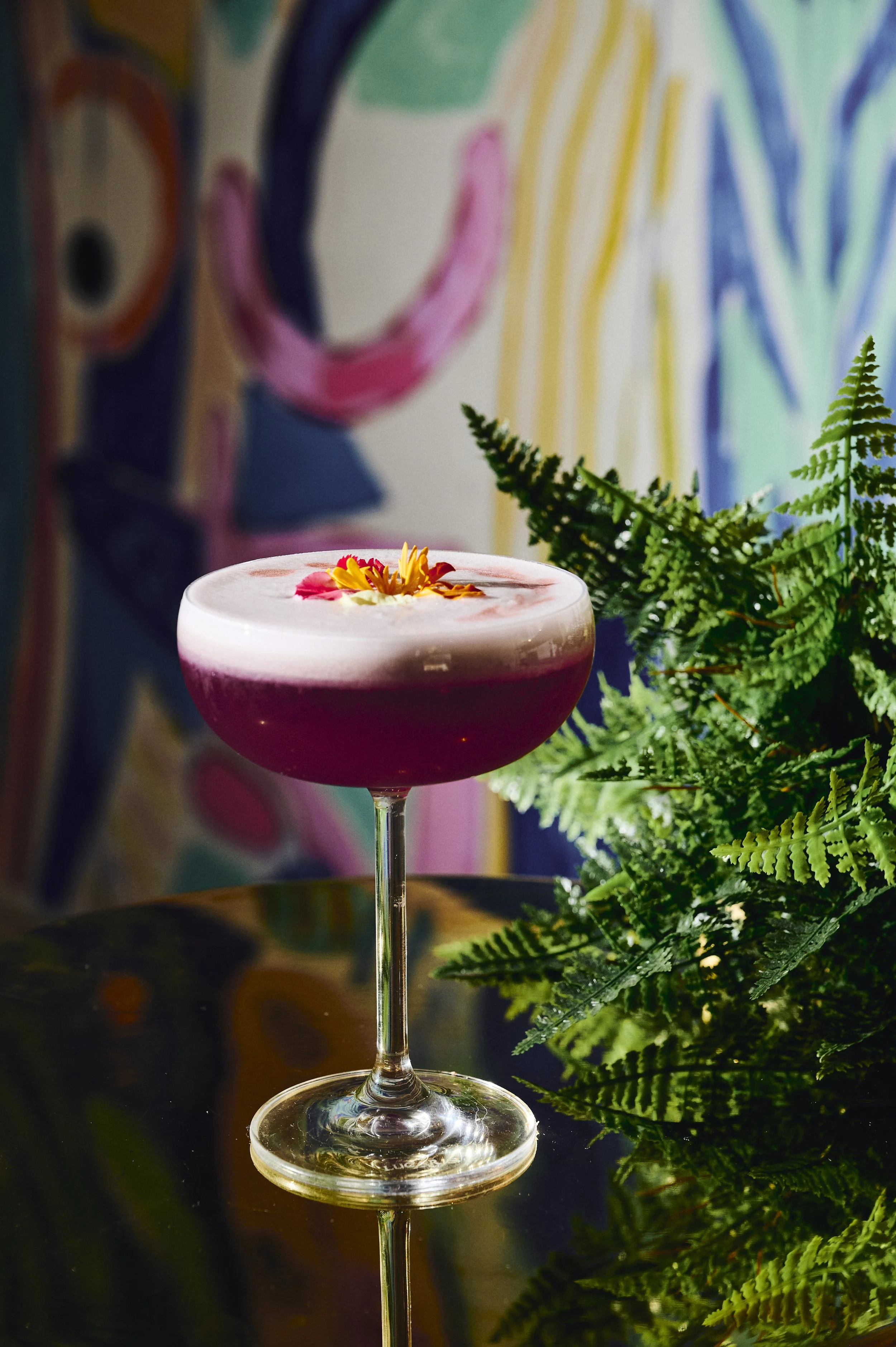 A cocktail in a coupe glass garnished with edible flowers, placed on a reflective surface with green fern leaves nearby, against a colorful, abstract background.