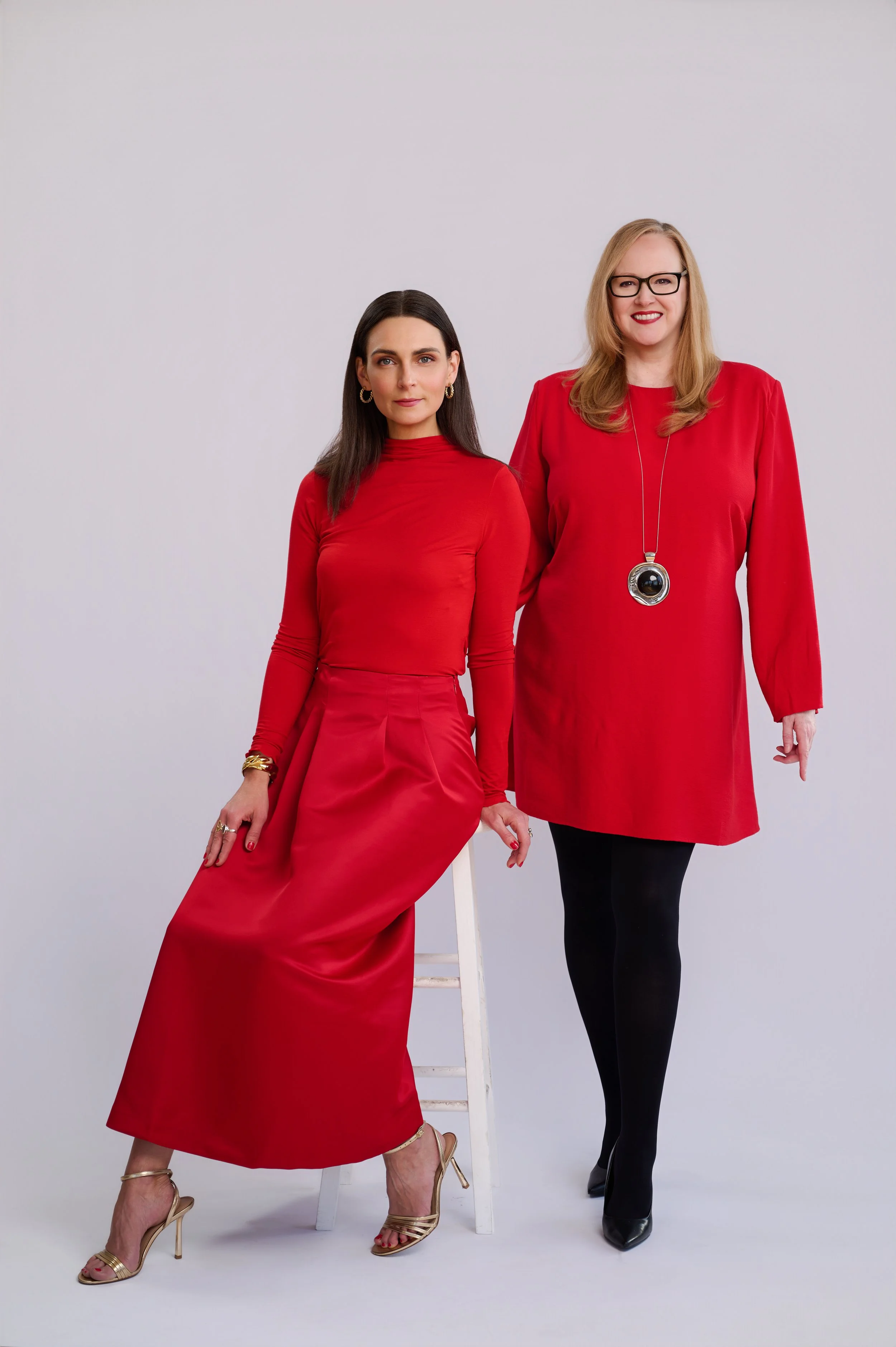 Two women, one seated on a white stool and the other standing, both wearing red dresses and black tights, posing against a plain white background.