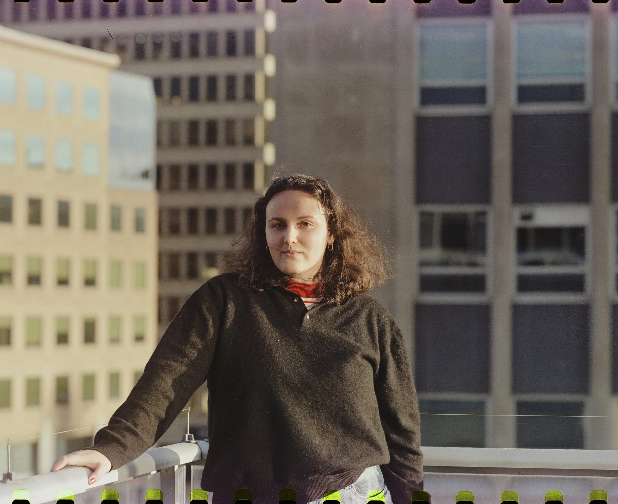 A young woman with dark, curly hair stands on a balcony in an urban setting, wearing a black sweater with a red and white striped shirt underneath. She is leaning with one arm on the railing, with tall buildings in the background.