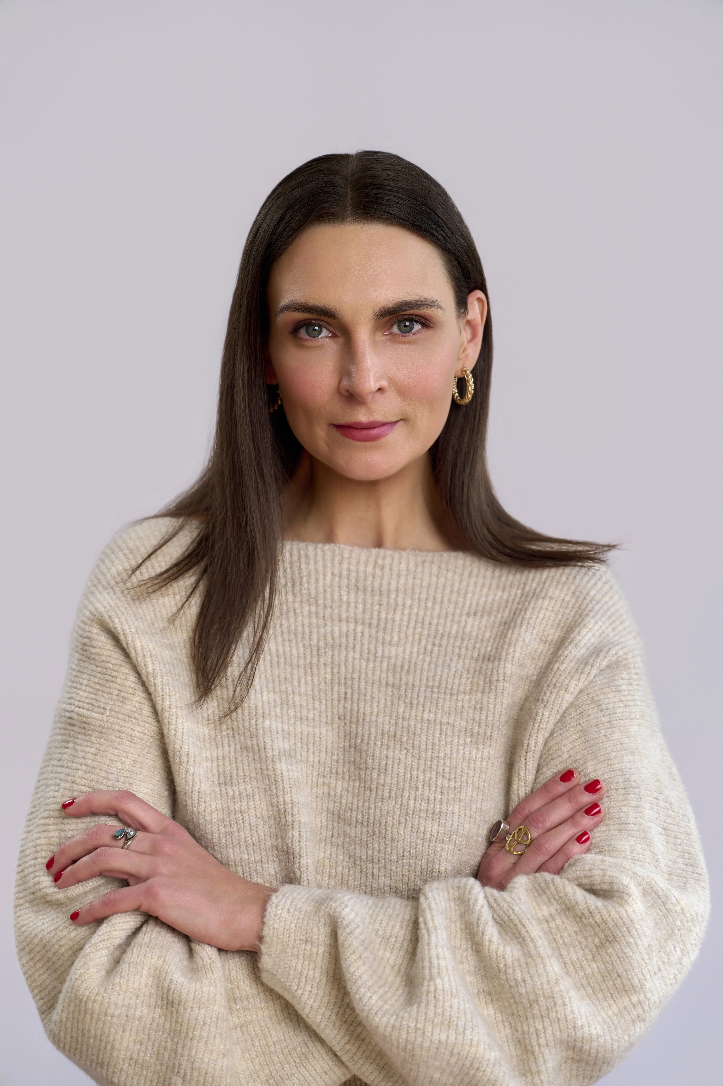 A woman with long brown hair wearing a beige sweater, standing against a light gray background, smiling with arms crossed.