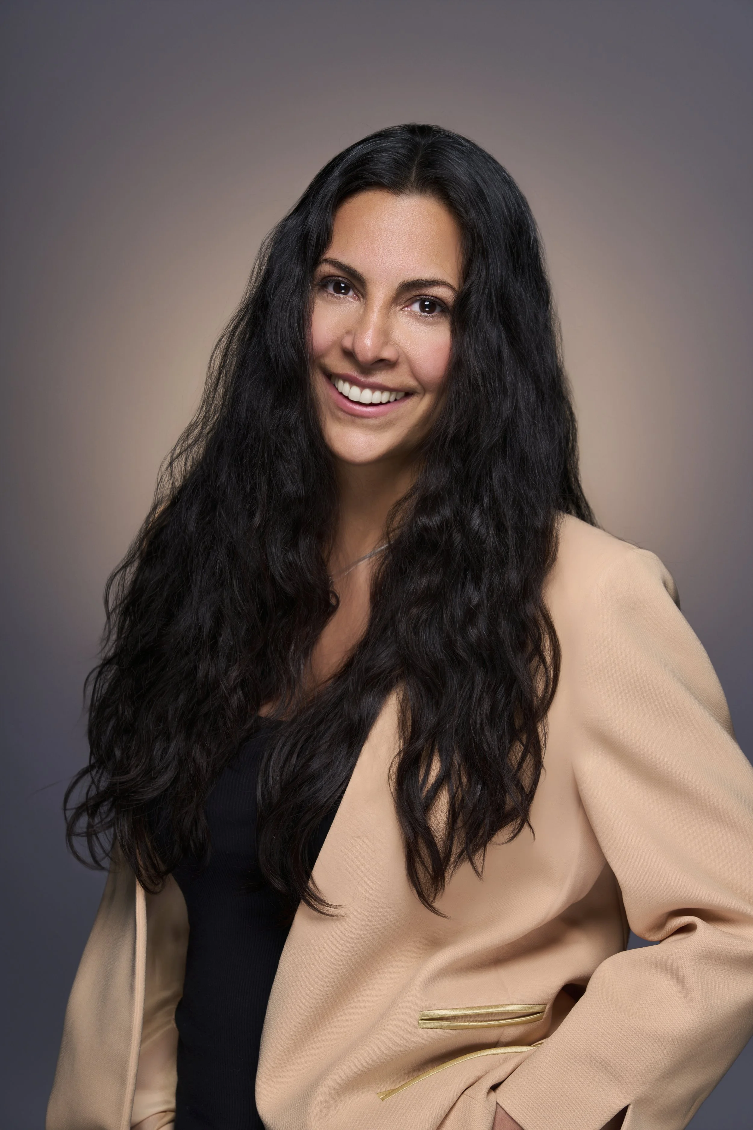 A woman with long, wavy black hair smiling at the camera, wearing a tan blazer over a black top, against a neutral background.