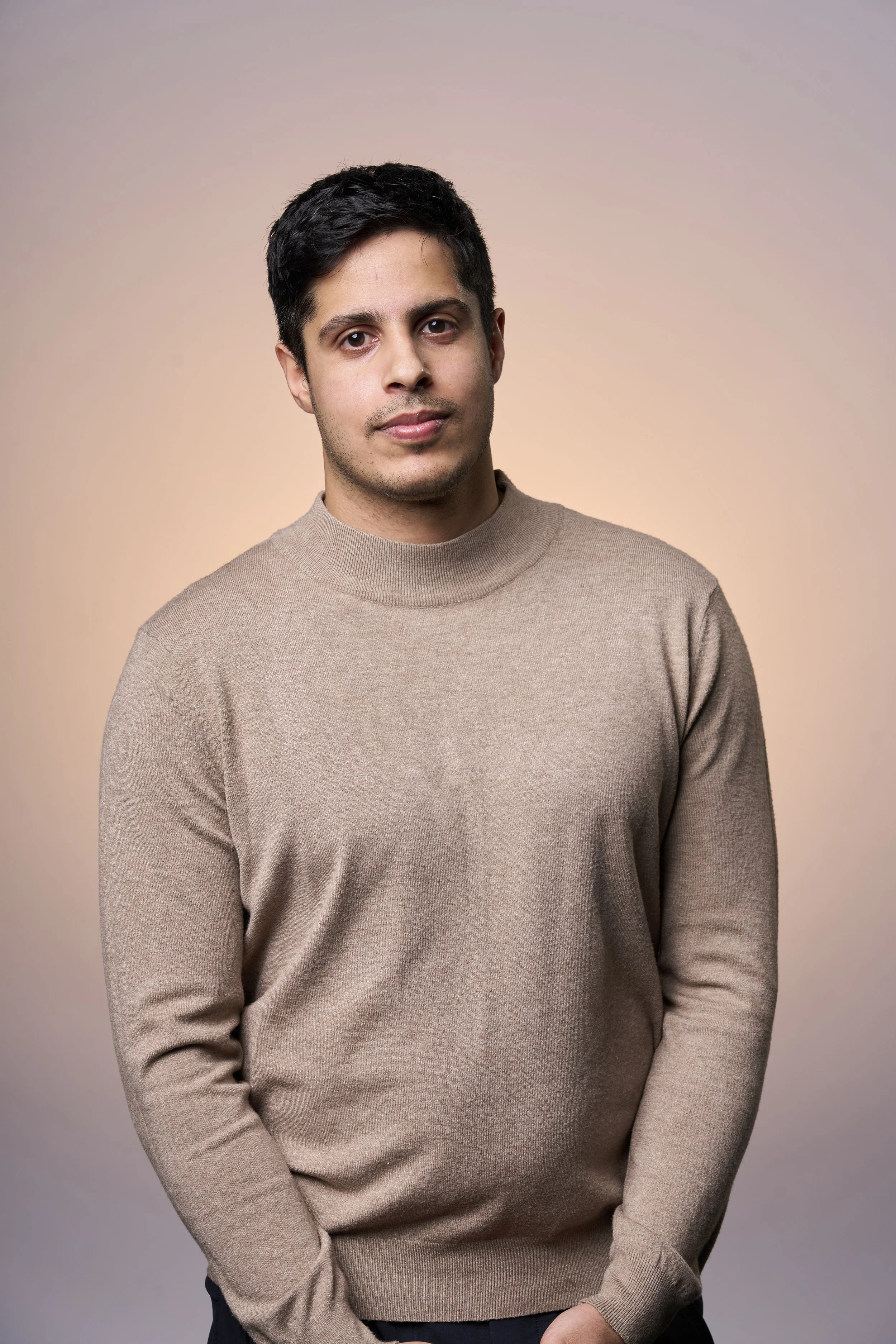 A portrait of a young man with dark hair, wearing a beige sweater, standing against a gradient background.