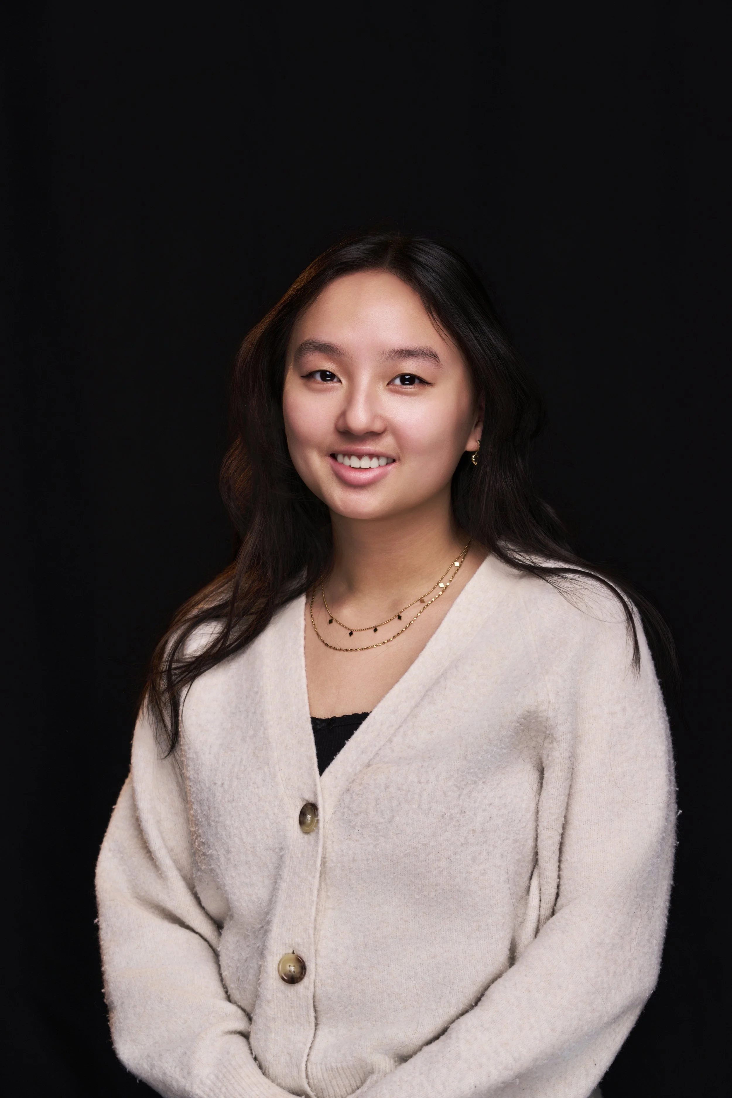 Portrait of a young woman with long dark hair, smiling, wearing a cream cardigan, layered necklaces, and earrings against a black background.
