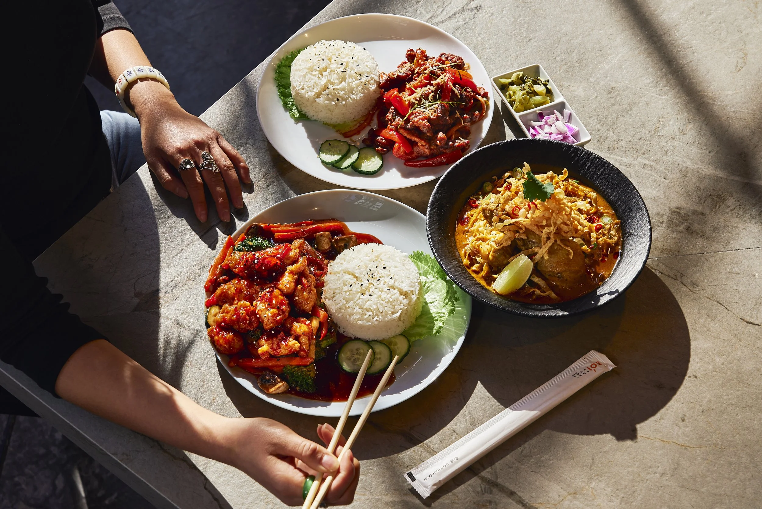 A table with Asian cuisine dishes including stir-fried vegetables with chicken, rice, savory curry with vegetables, and small side dishes of pickled vegetables. A person is using chopsticks to pick up food.