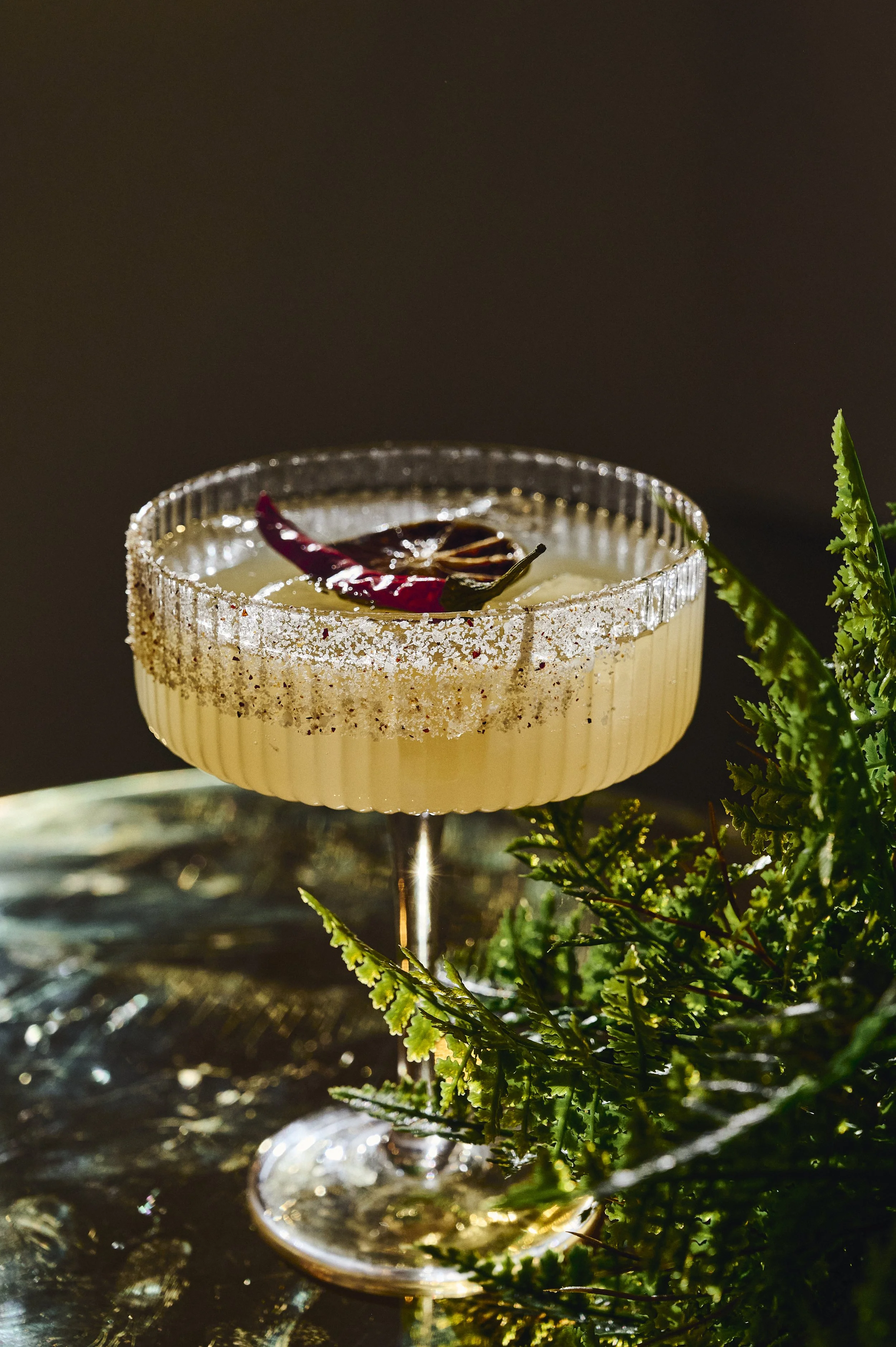 A cocktail in a ribbed glass with a salted rim, garnished with a dried red chili and a star anise, placed on a reflective surface surrounded by green foliage.